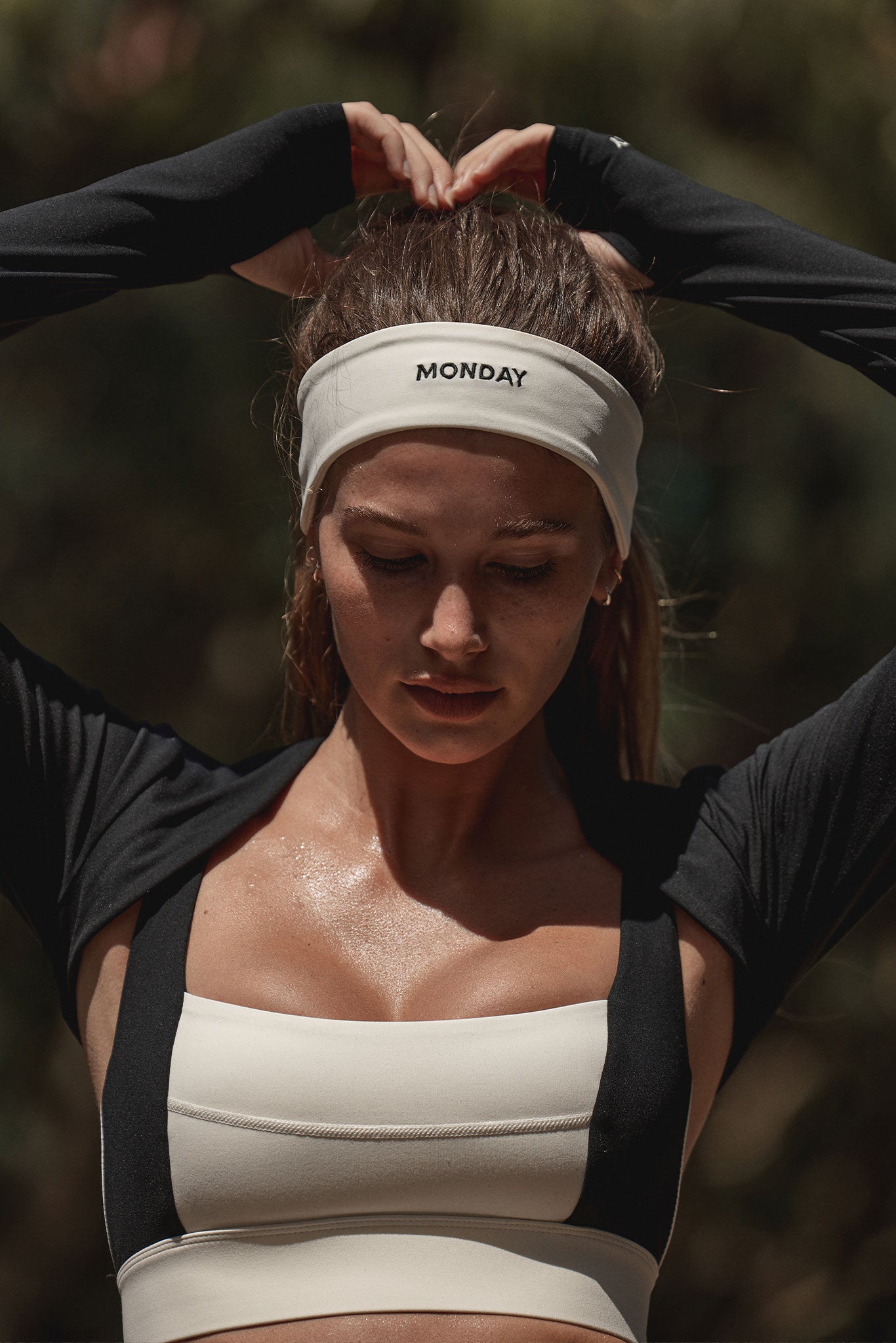 Woman wearing a white headband labeled MONDAY and cream sports bra, hands raised above head, looking down.