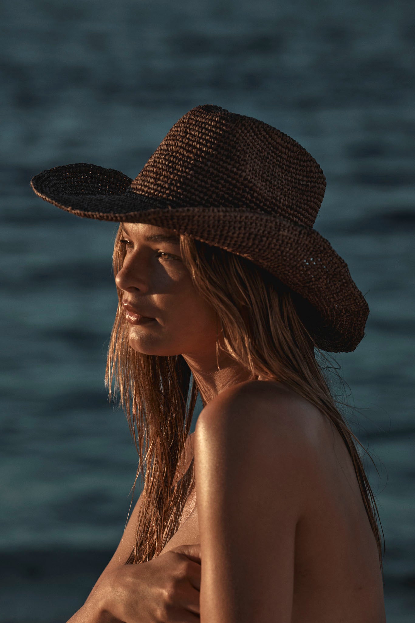 Woman wearing a woven straw hat looking toward the sea at sunset, bare shoulders with arm across chest.