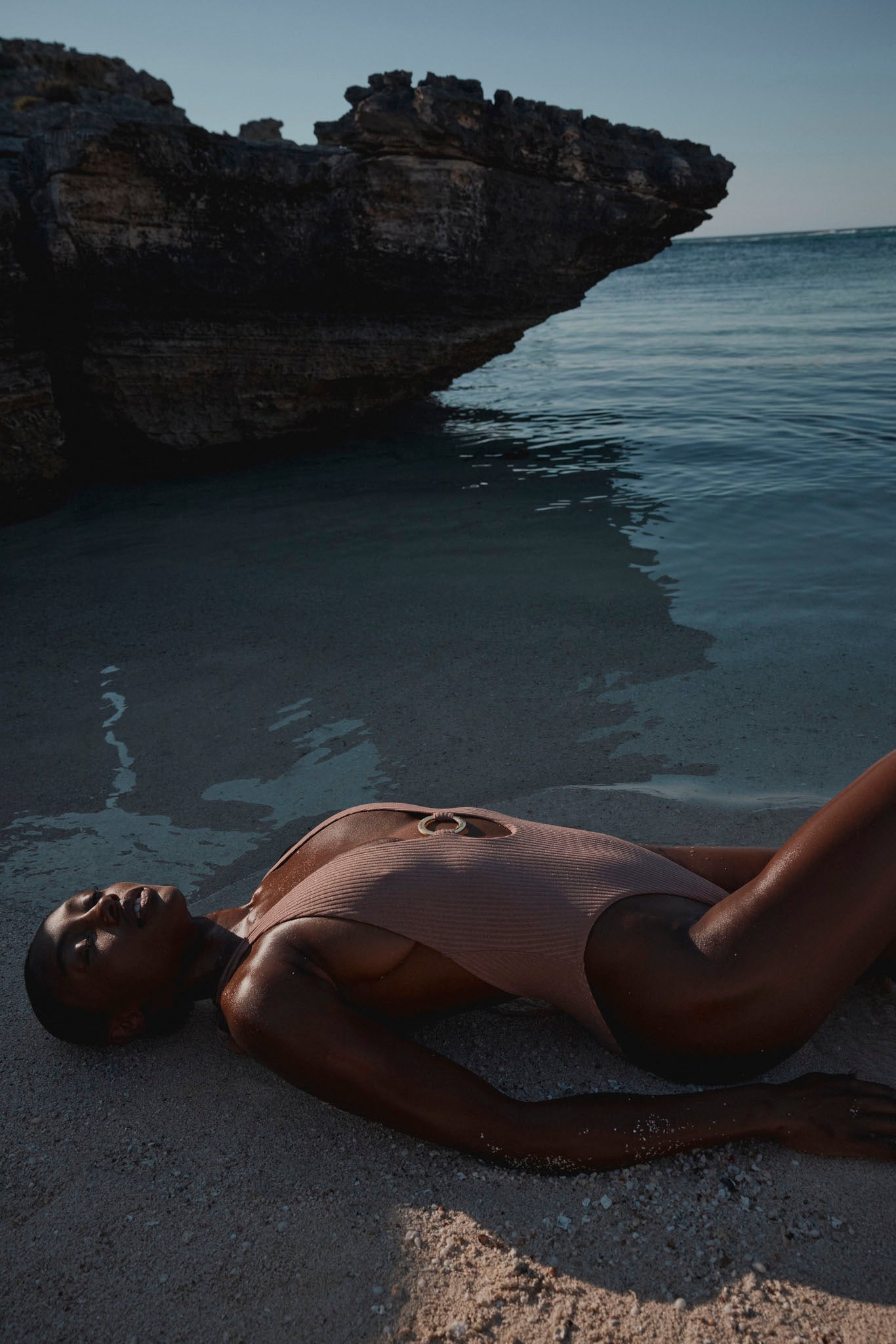 Person reclining on sandy shore at the water's edge in a one-piece swimsuit beneath a rocky overhang and calm sea.