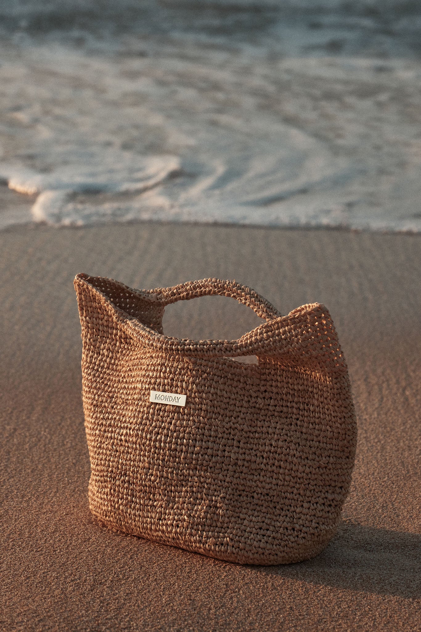 Woven straw beach tote labeled MONDAY resting on wet sand near small incoming waves.
