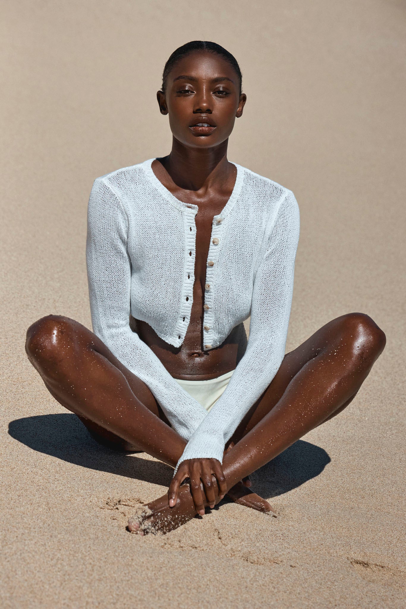 Woman seated on sandy beach wearing a cropped white knit cardigan and bikini bottoms, legs crossed, hands clasped over feet.