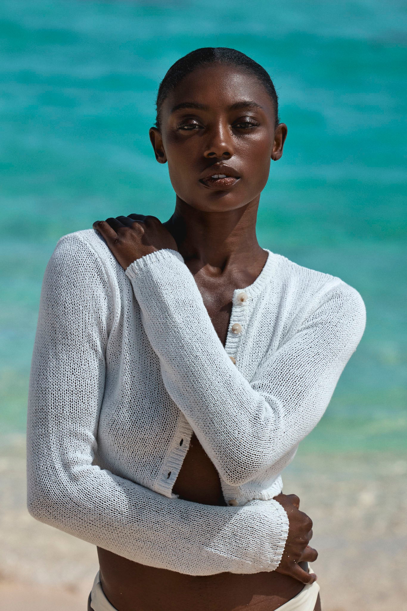 Woman in a white knit cardigan with arms crossed, standing on a beach with turquoise water