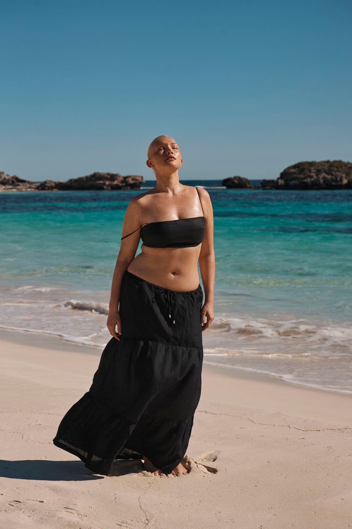 Bareheaded model standing barefoot on sunlit beach in black bandeau top and flowing black skirt, eyes closed toward sky.