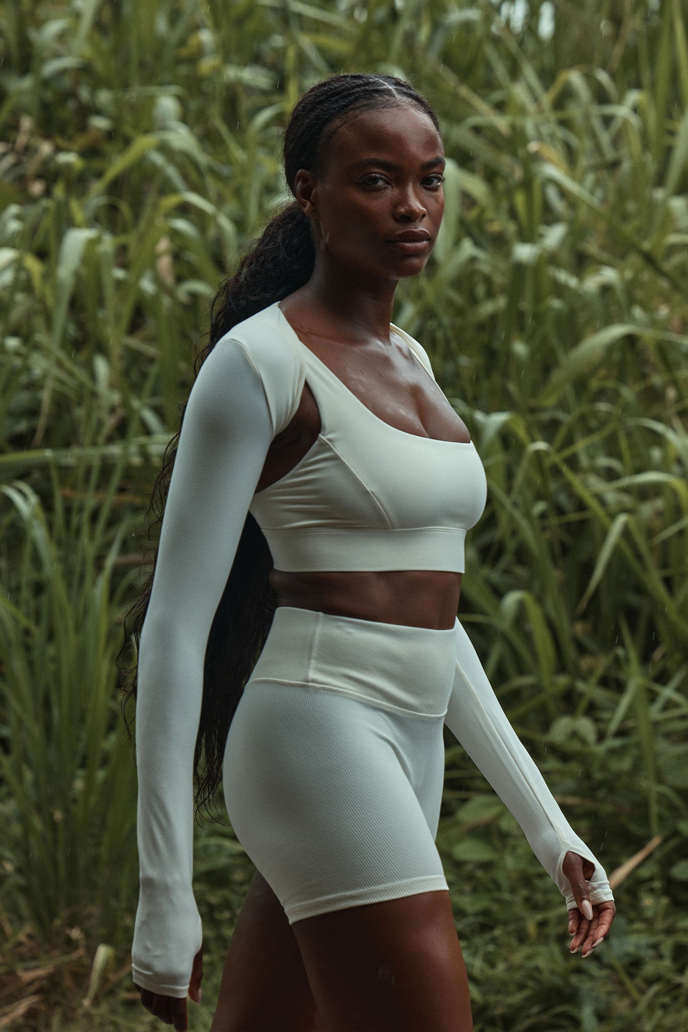 Woman with long braided hair wearing white fitted sport top and shorts standing in tall green grass