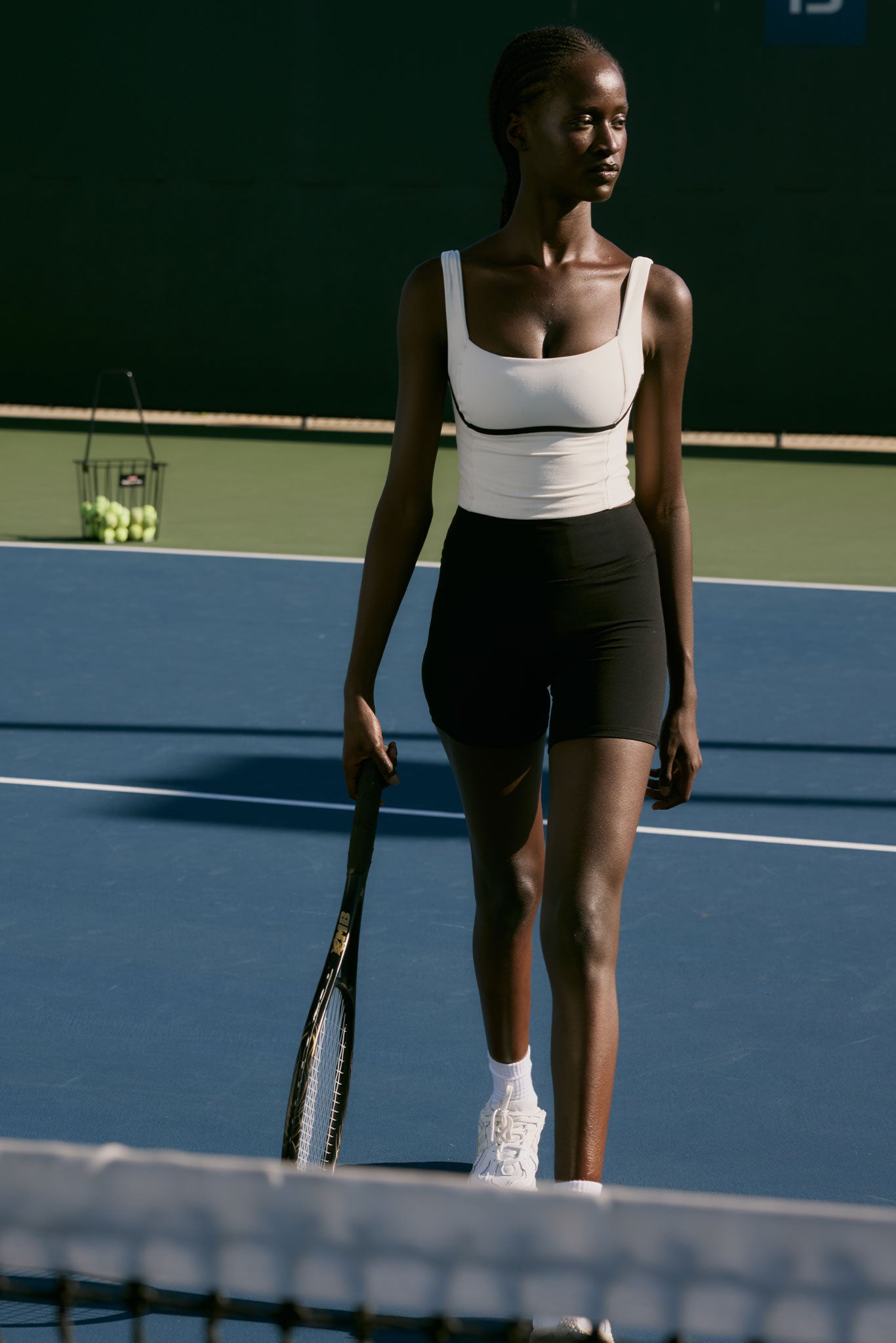 Female tennis player standing on a court holding a racket, wearing white top, black shorts; basket of tennis balls behind her