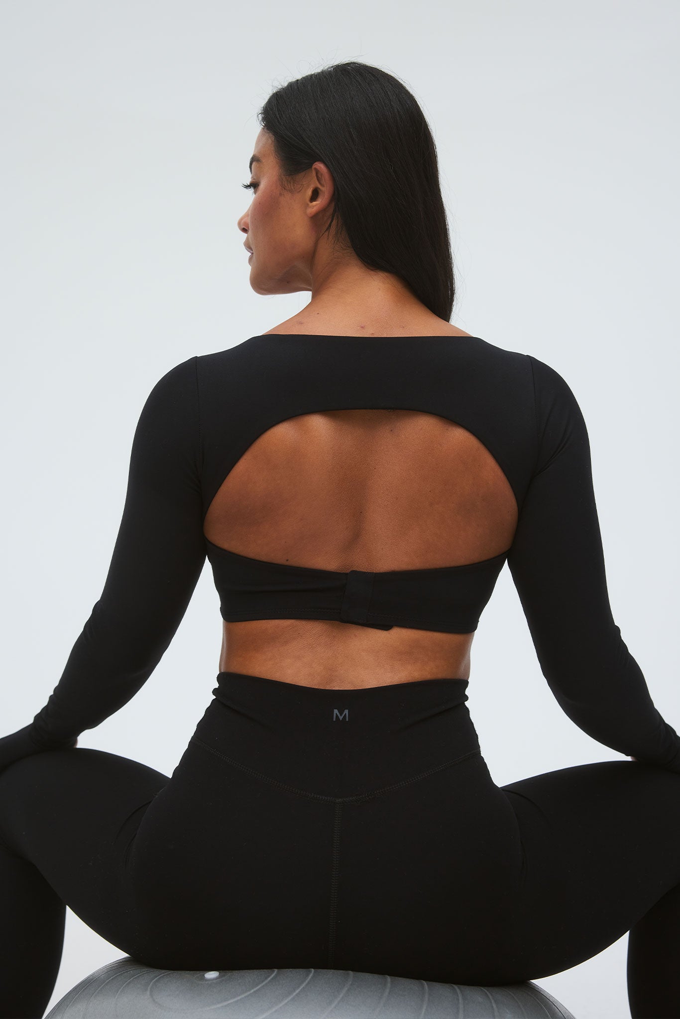 Back view of woman on exercise ball wearing black long-sleeve crop top with oval cutout and high-waist leggings marked 'M'.