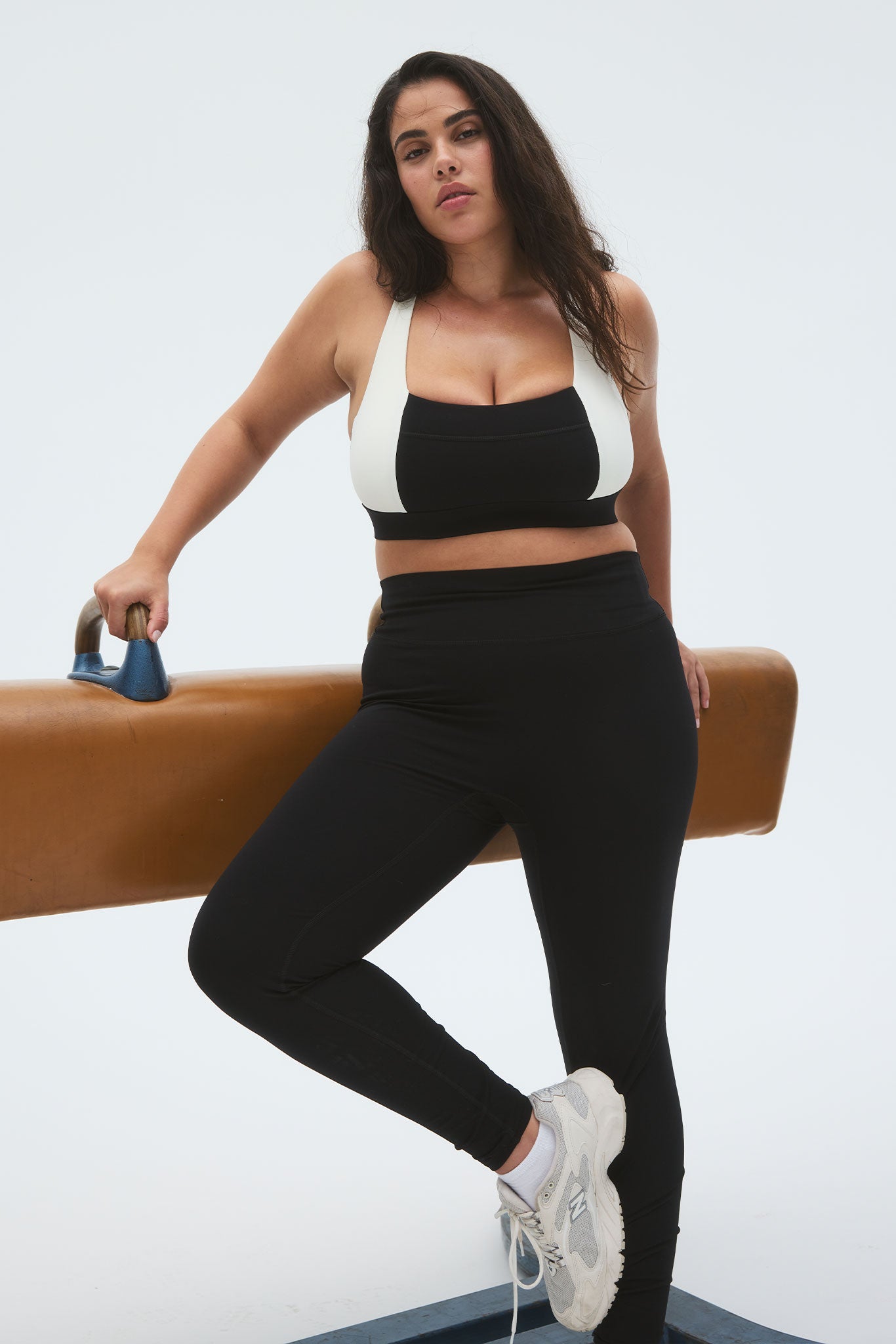 Plus-size woman in black-and-white sports bra and black leggings leaning on a brown pommel horse, wearing white sneakers