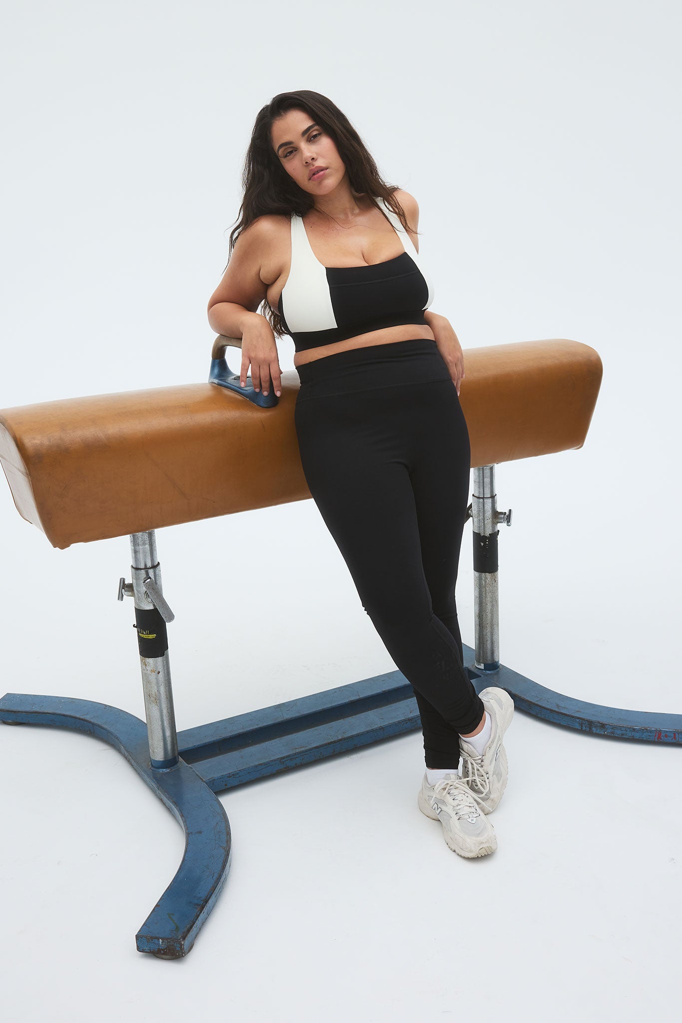 Woman leaning against a leather pommel horse wearing black-and-white sports bra, high-waist black leggings and white sneakers.