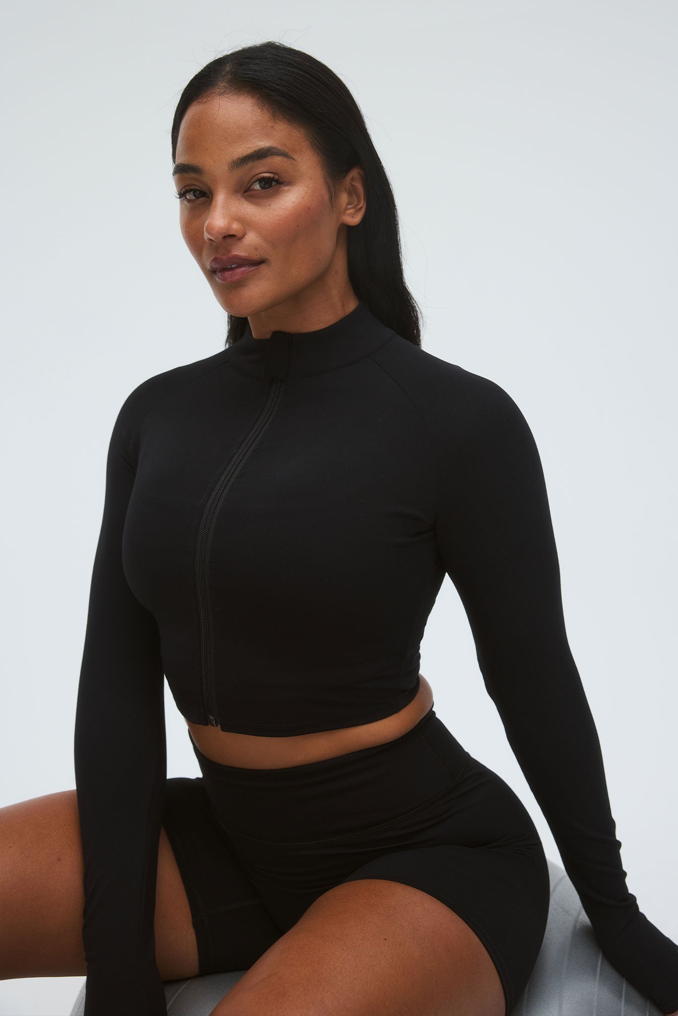 Female model seated wearing black zip-front long-sleeve crop top and black high-waisted bike shorts.