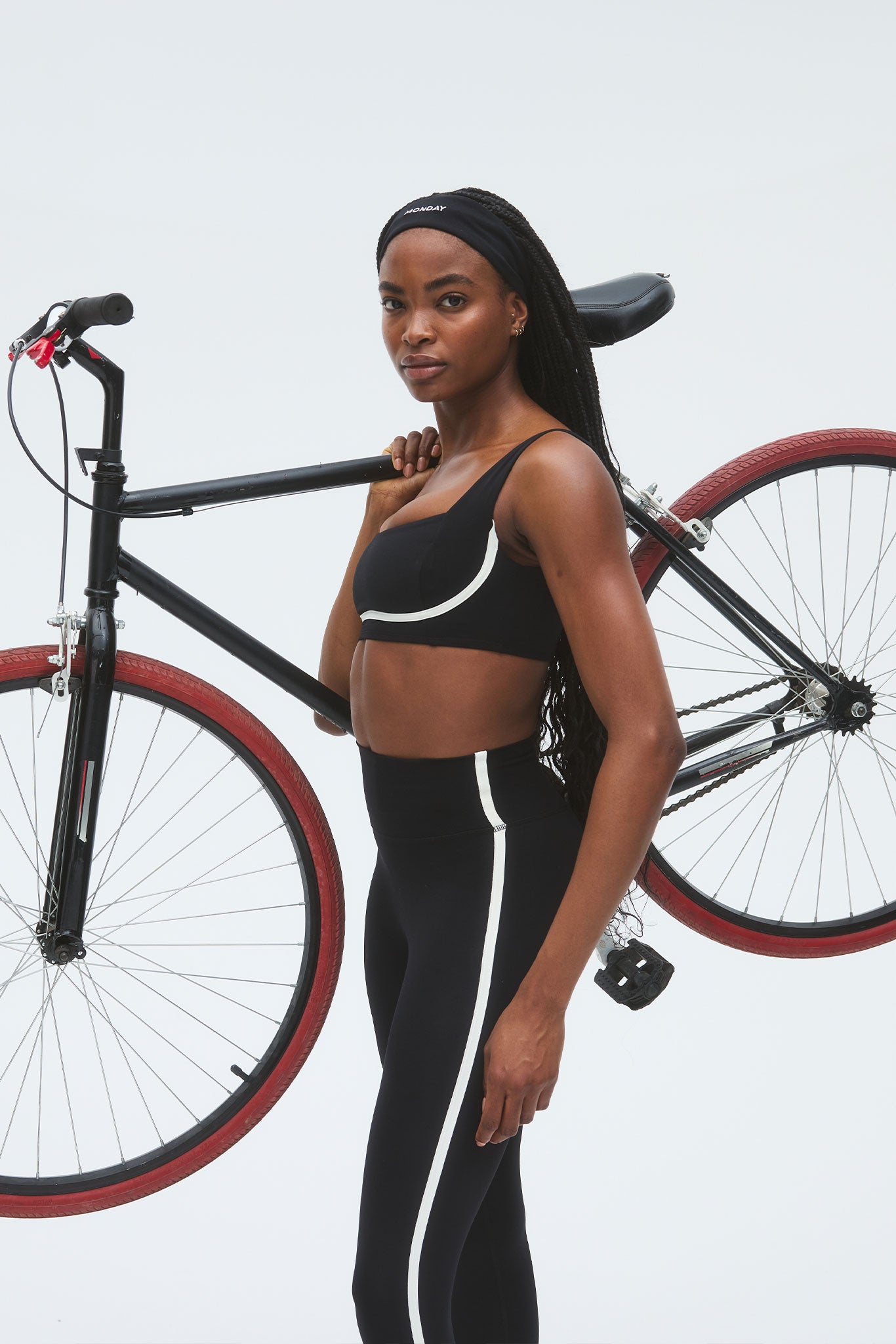 Woman carrying a black bicycle on her shoulder, wearing black sports bra and leggings with a white stripe; headband reads MONDAY