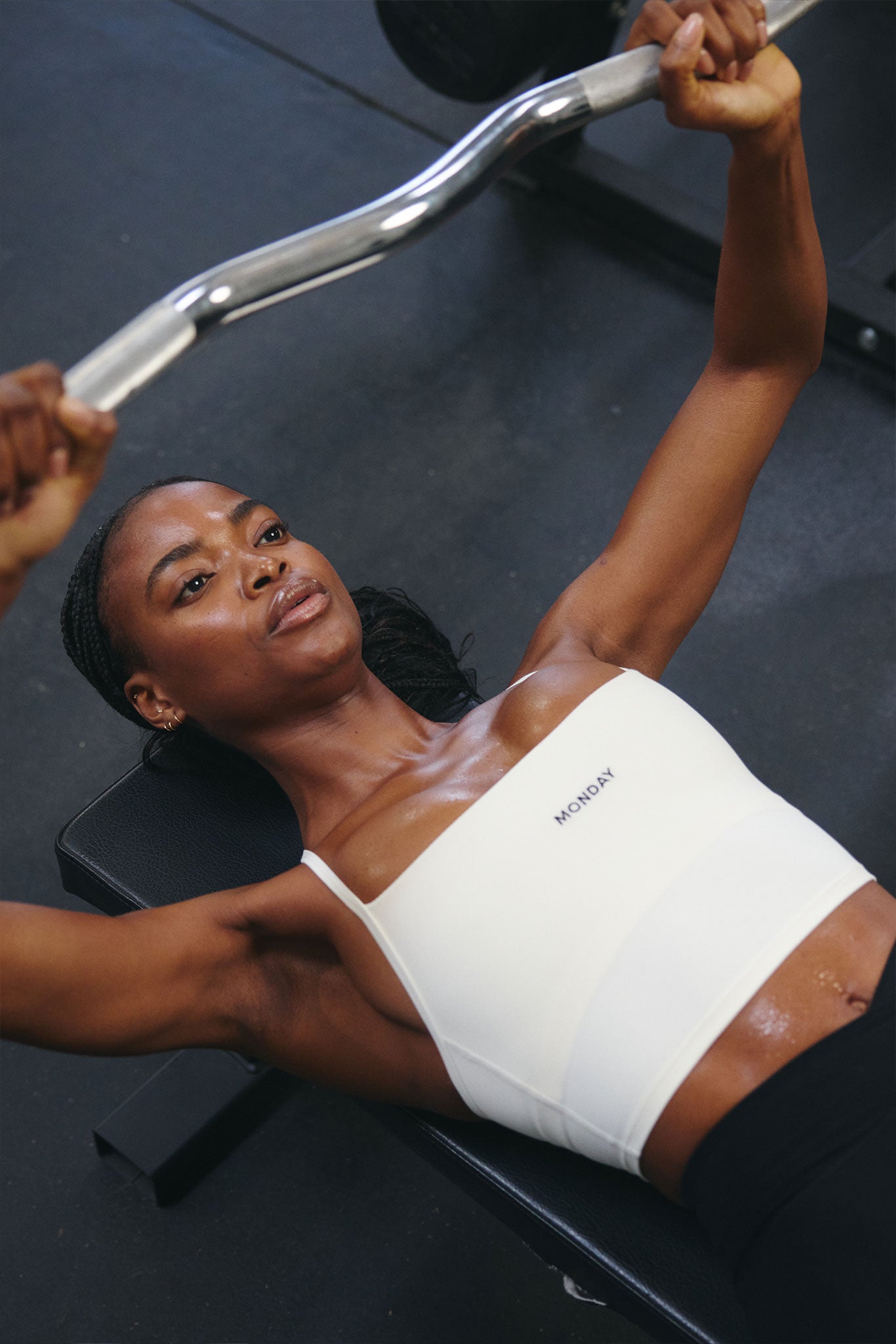 Woman lying on bench pressing a curved barbell, wearing a white crop top labeled MONDAY.