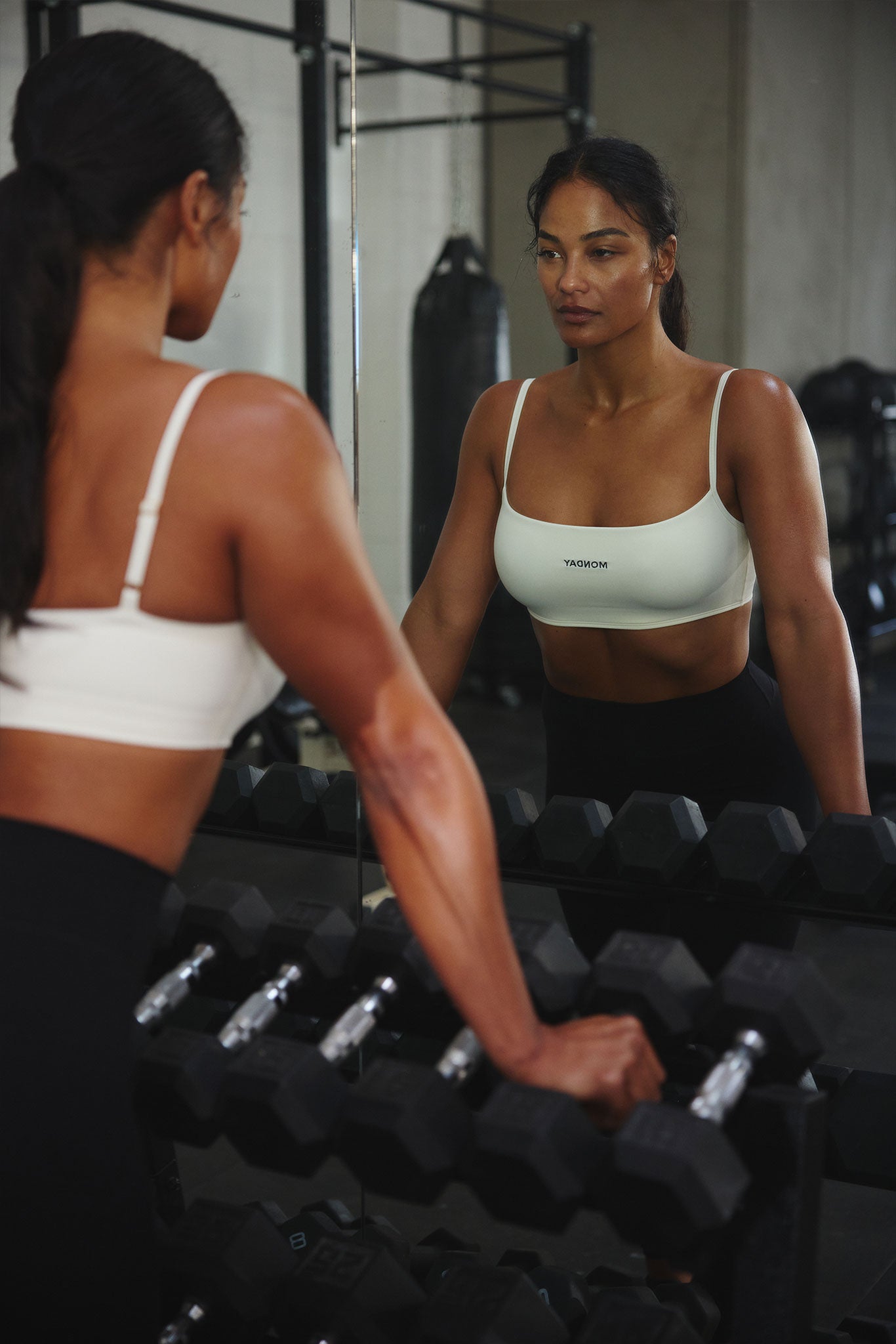 Athletic woman gazing at her reflection in a gym mirror, hands on a dumbbell rack, wearing a white sports bra labeled MONDAY
