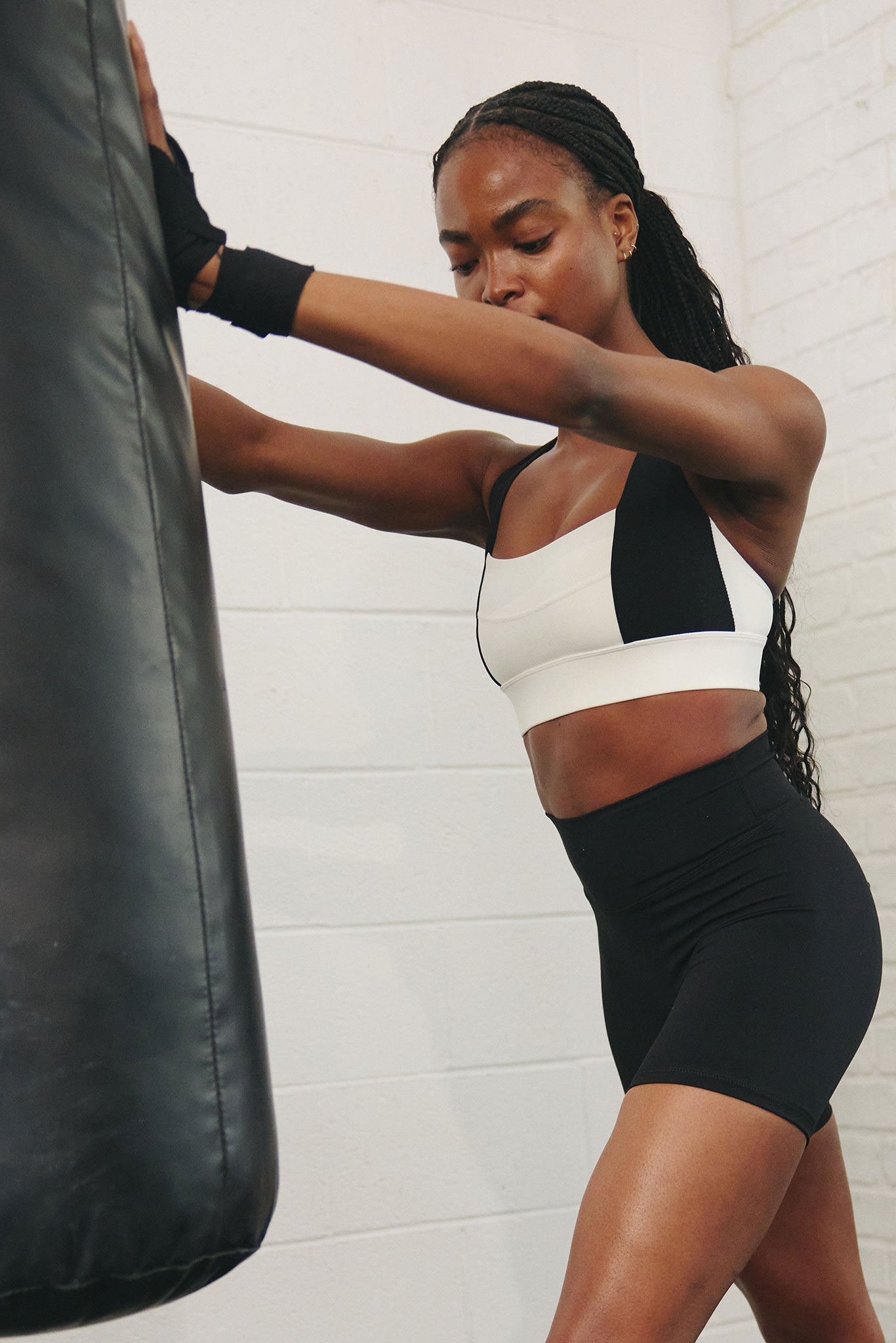 Woman in sports bra and high-waist shorts leaning on a heavy punching bag, preparing to box.