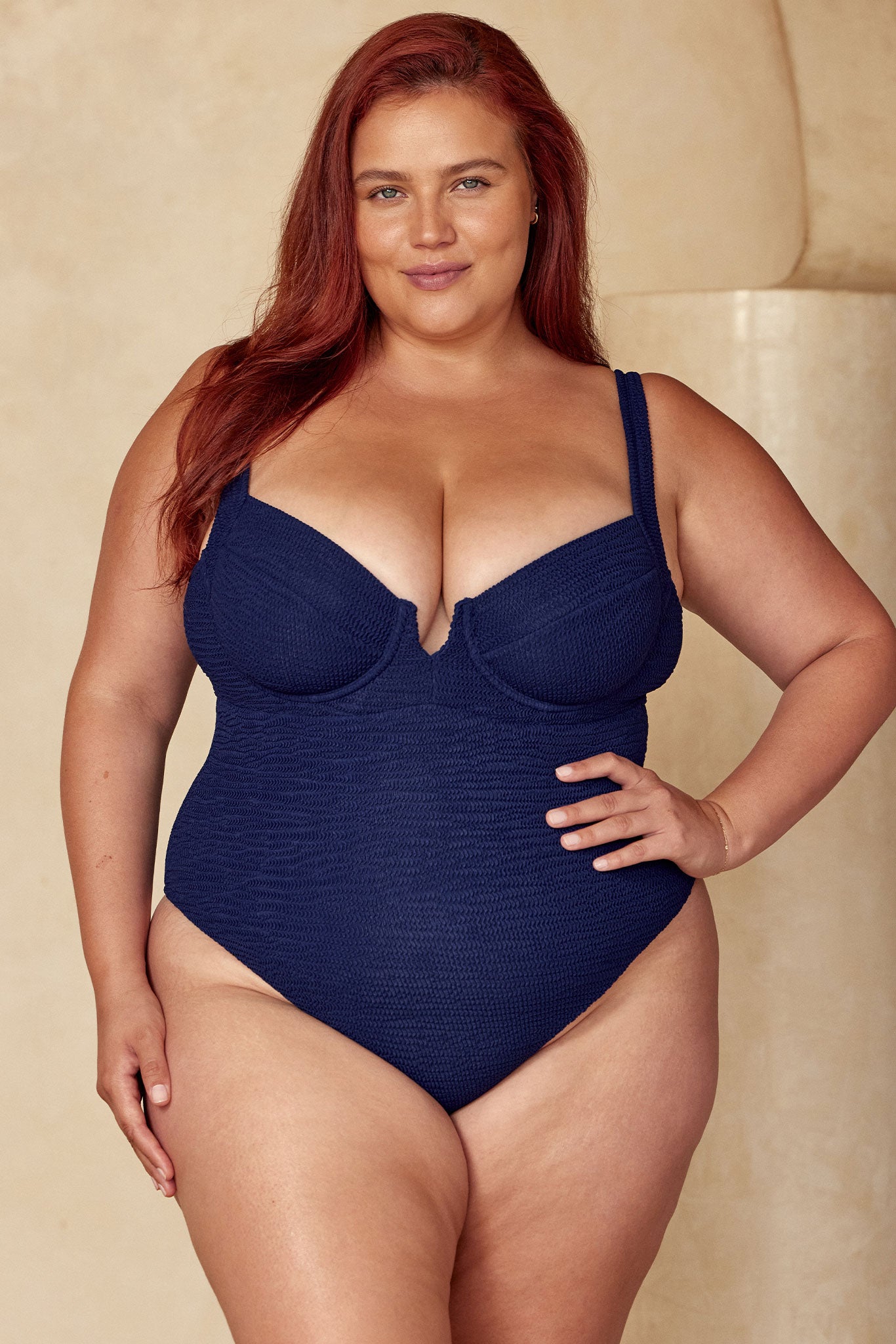 Plus-size model wearing navy textured underwire one-piece swimsuit with shoulder straps, hand on hip, facing camera.
