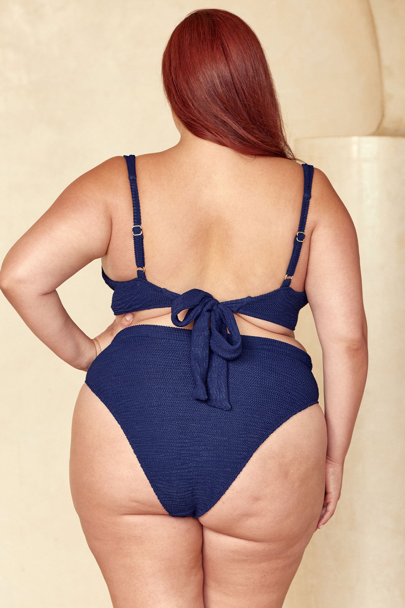 Back view of a plus-size model wearing a navy textured tie-back bikini with high-waist bottoms