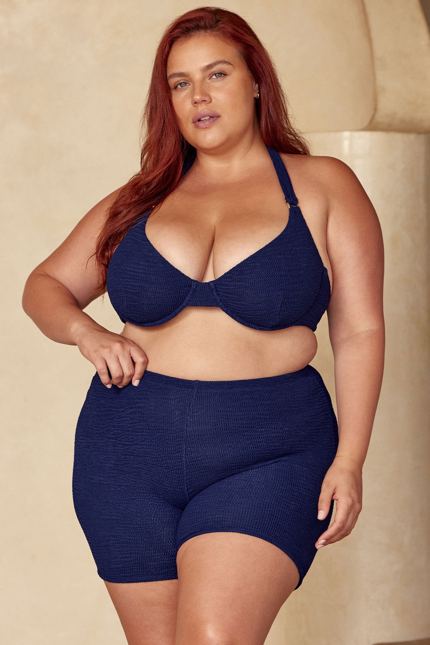 Plus-size woman standing in navy textured bikini top and high-waisted swim shorts, hand on waist, neutral background