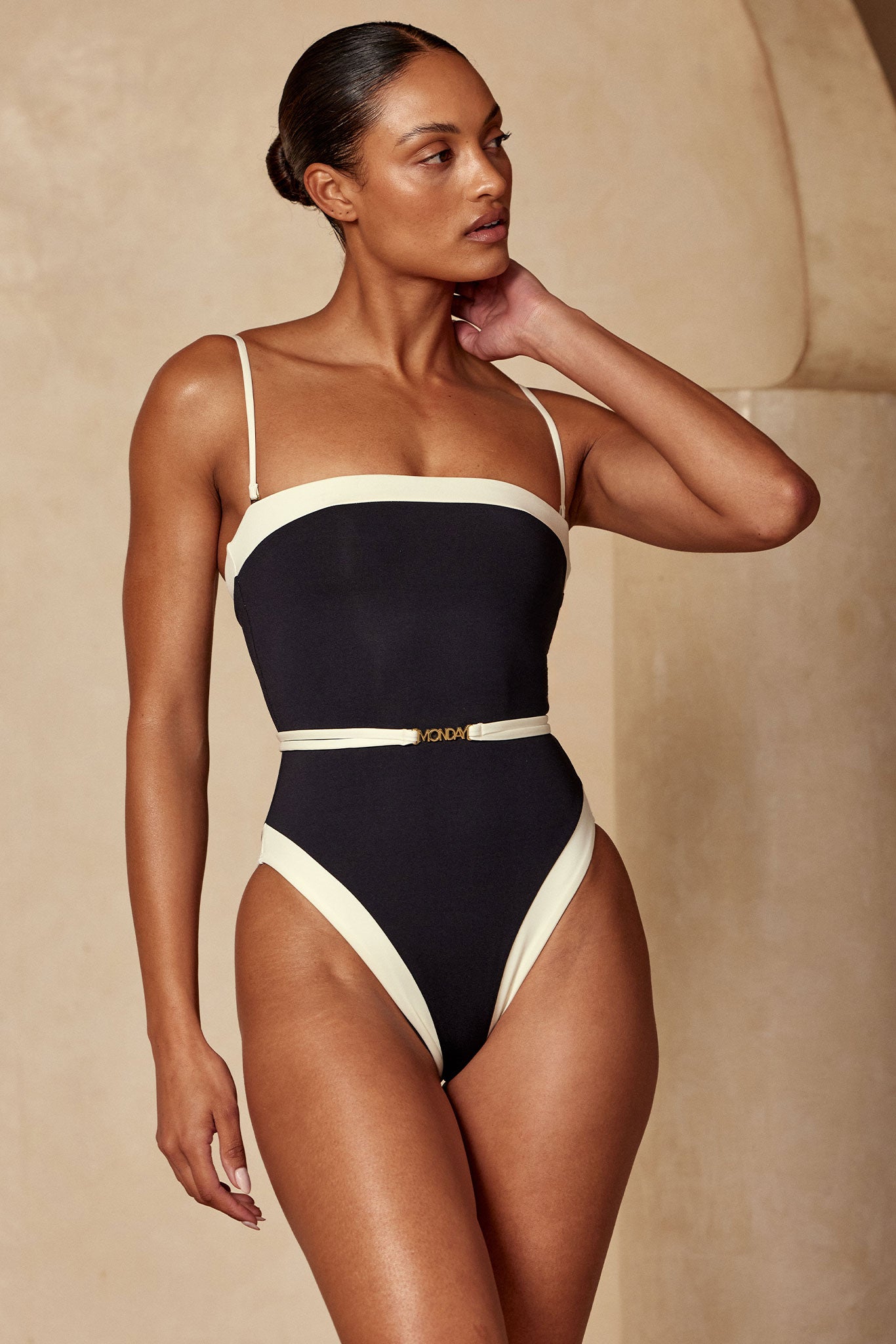 Model wearing black one-piece swimsuit with cream trim and skinny belt buckle reading MONDAY.