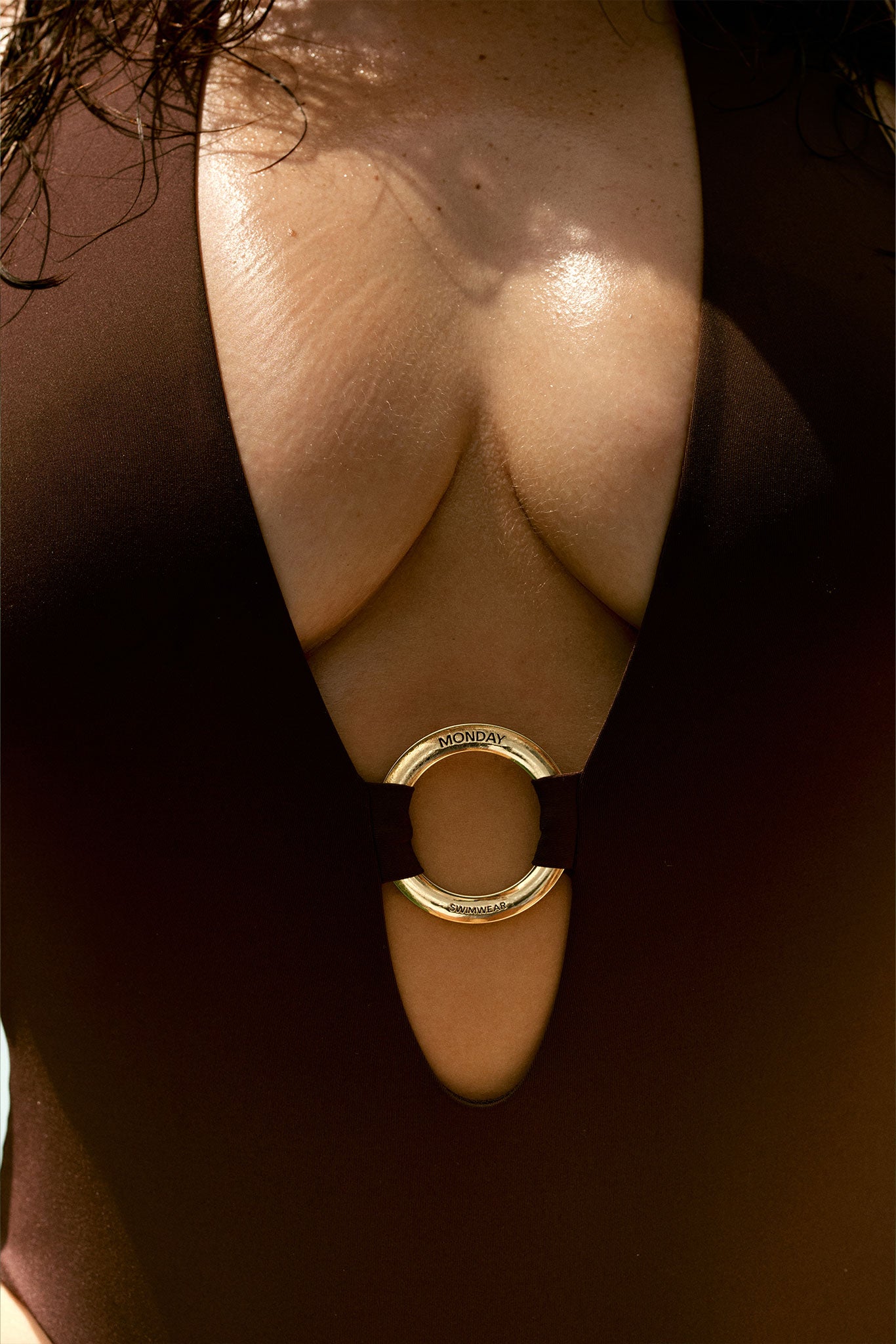 Close-up of swimsuit front with gold ring labeled MONDAY SWIMWEAR, showing neckline, fabric and cleavage.