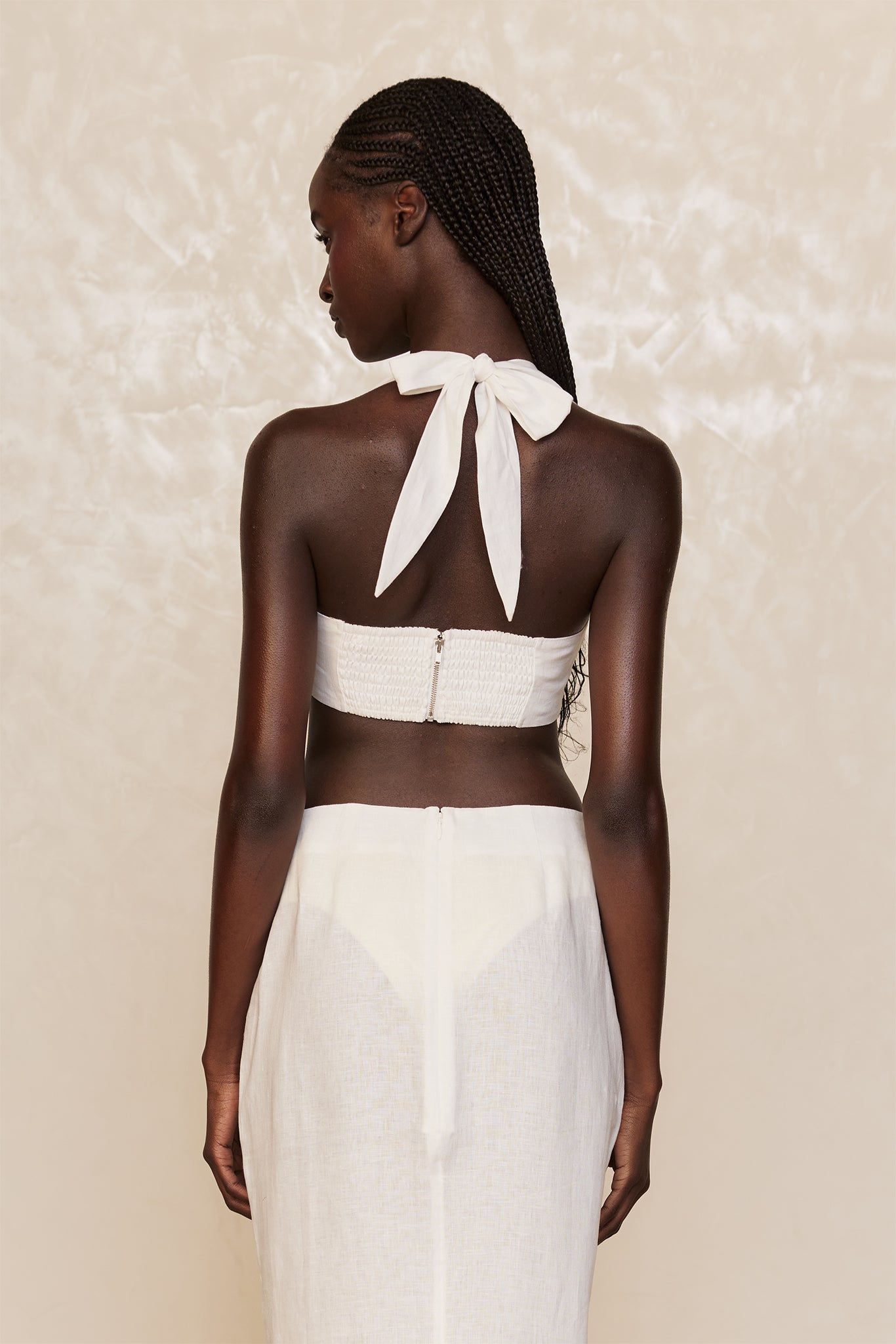 Back view of woman with long braided hair wearing a white halter top tied in a bow and a white linen skirt.