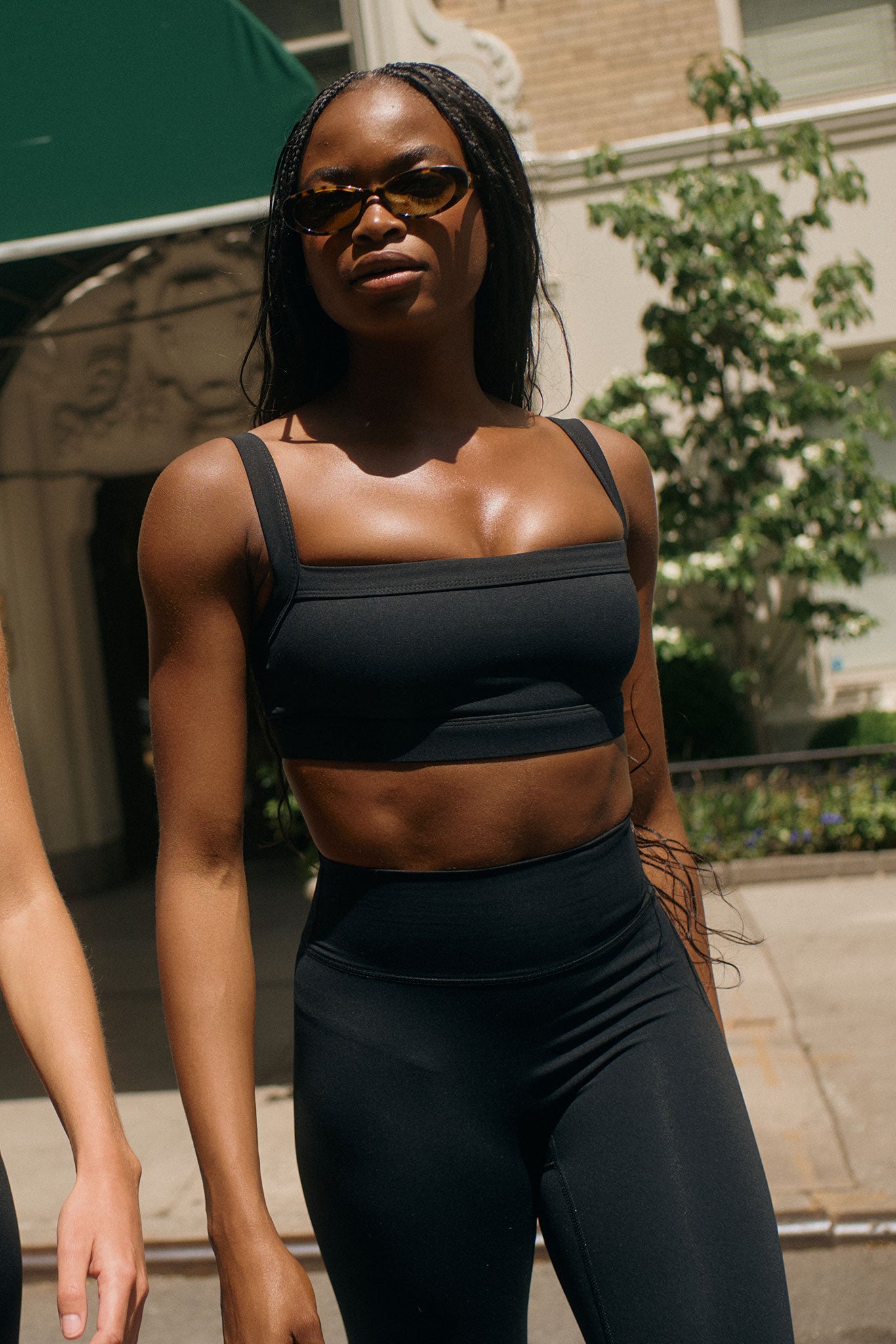 Black woman in black sports bra and high-waist leggings wearing sunglasses, walking outdoors on a sunlit city street.