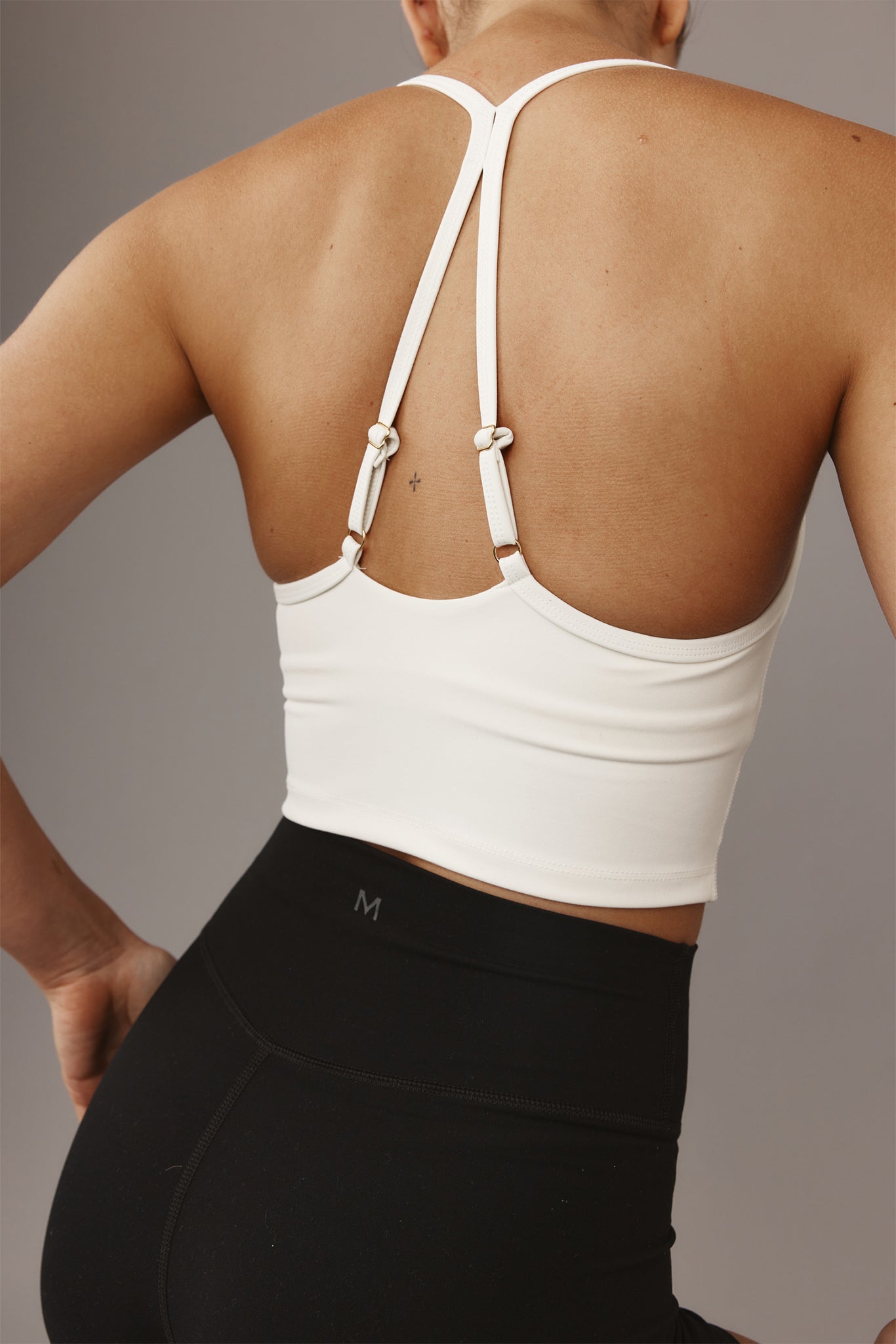 Woman's back wearing white strappy sports bra and black high-waist leggings; small 'M' logo on waistband.