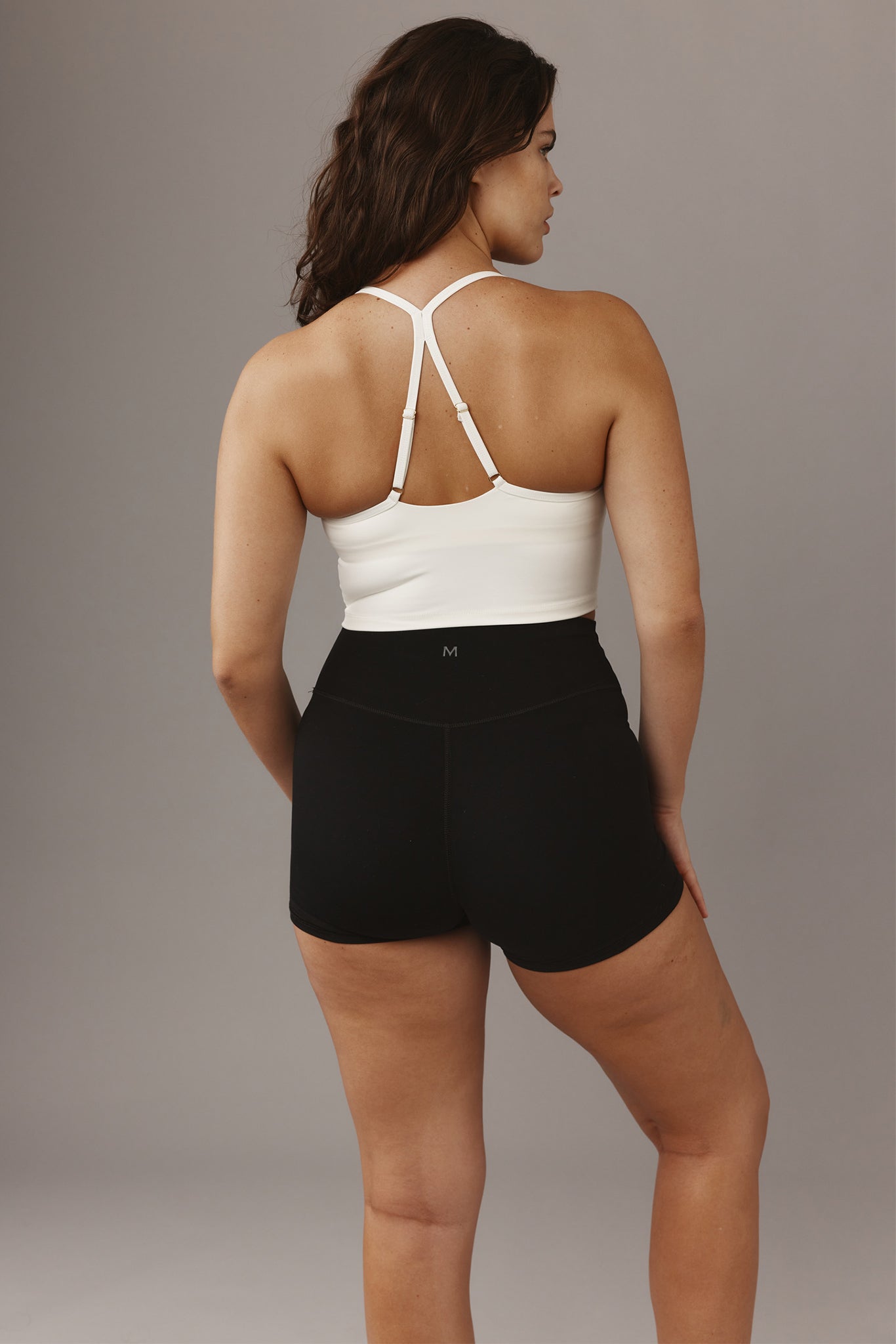 Back view of woman wearing a white strappy T-back sports bra and high-waist black shorts with small 'M' logo on waistband.