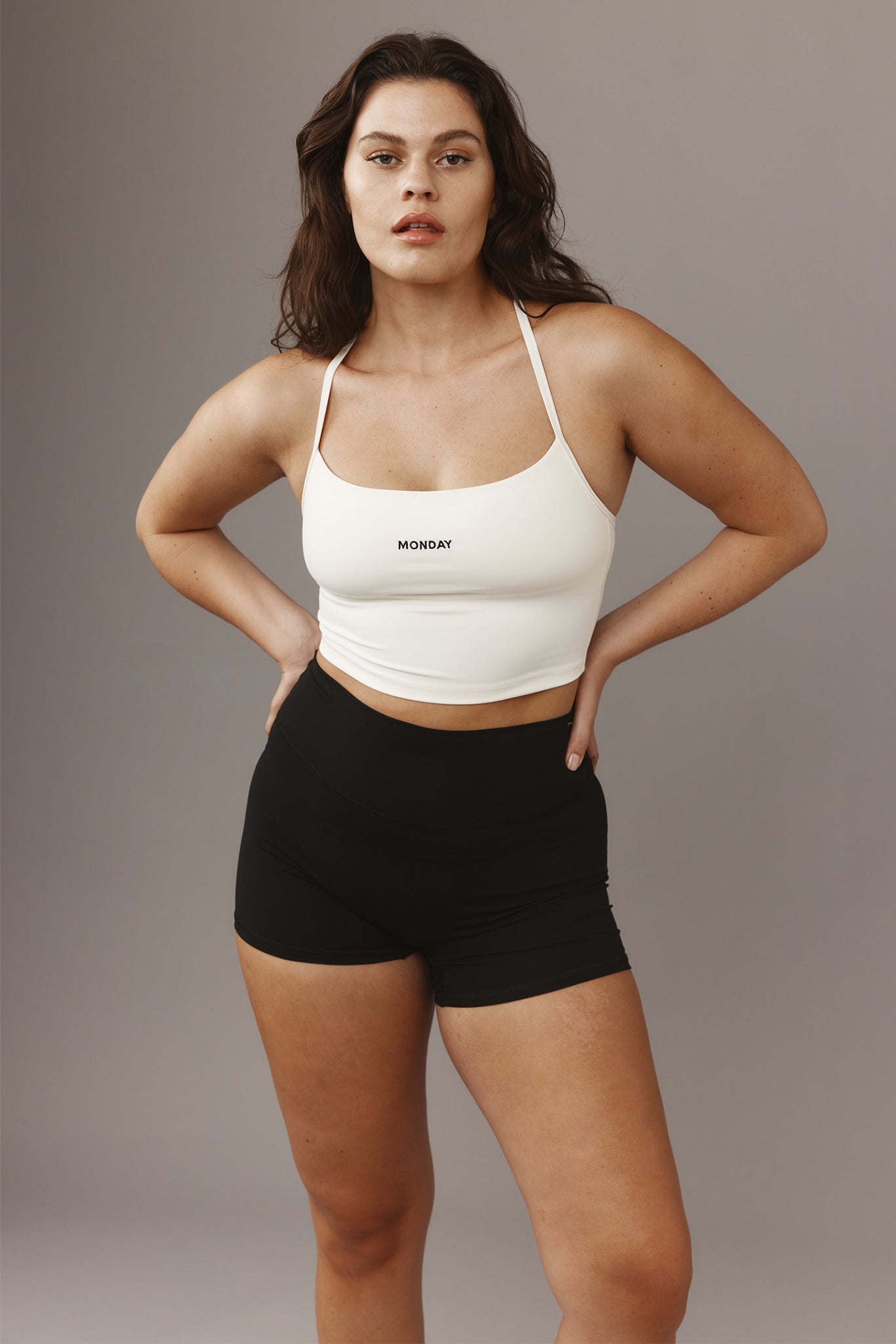 Woman standing with hands on hips wearing white cropped tank top reading 'MONDAY' and black high-waist shorts on gray background.