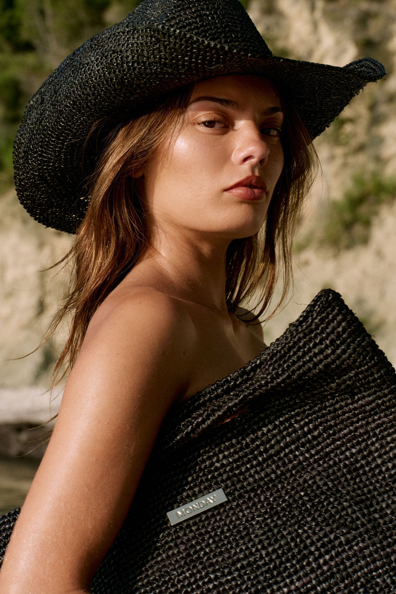 Woman wearing a black straw hat holding a woven black bag labeled MONDAY.