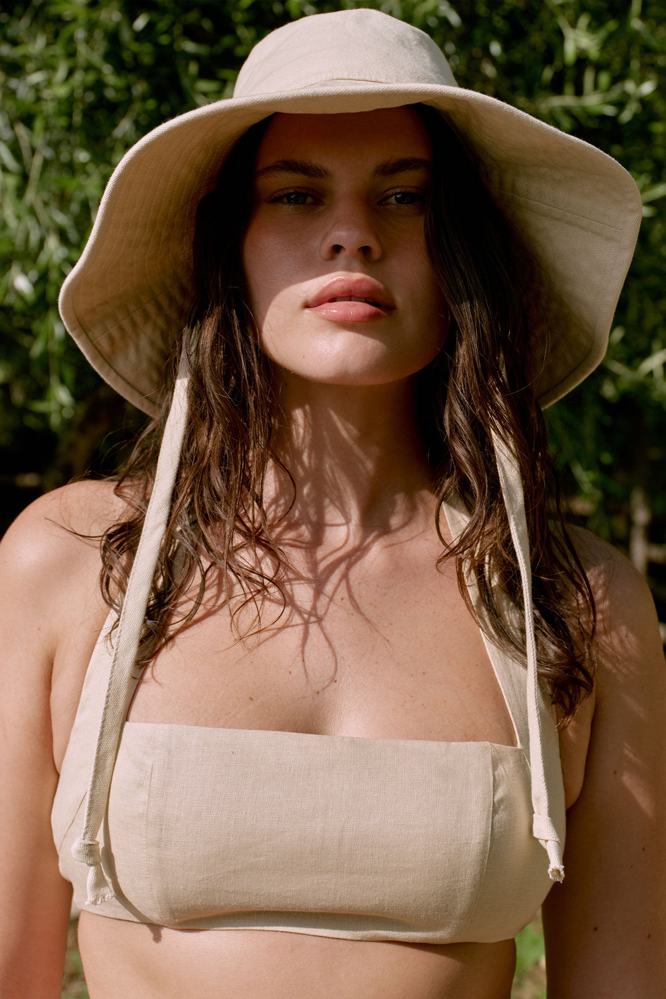 Close-up of a woman wearing a beige sun hat and linen bandeau top, with sunlit shadows across her face and chest.