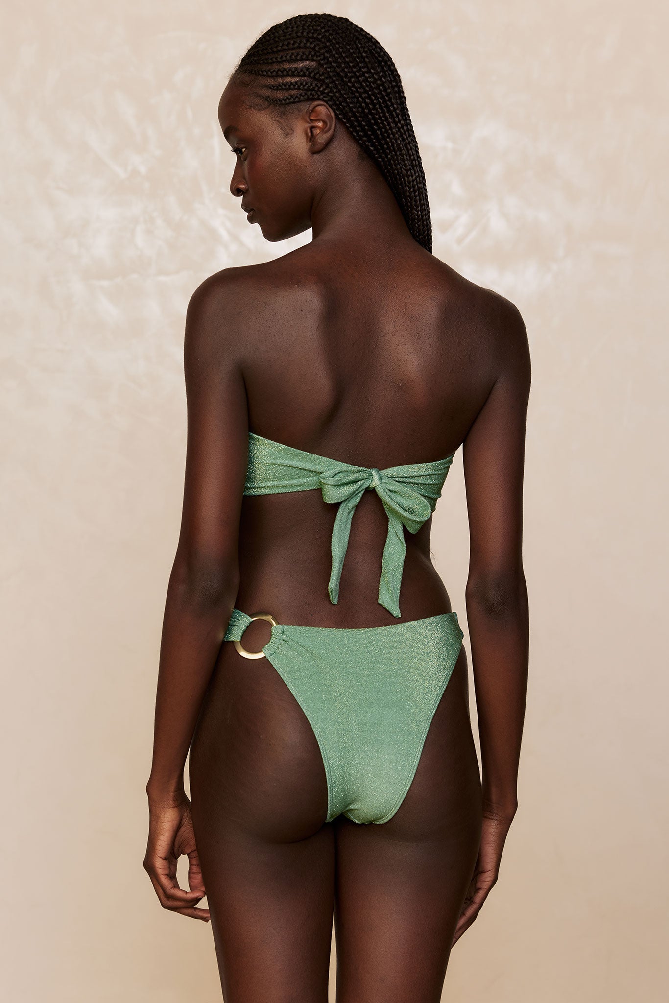 Back view of woman in metallic green bikini with tied bandeau top and ring-detail high-cut bottoms