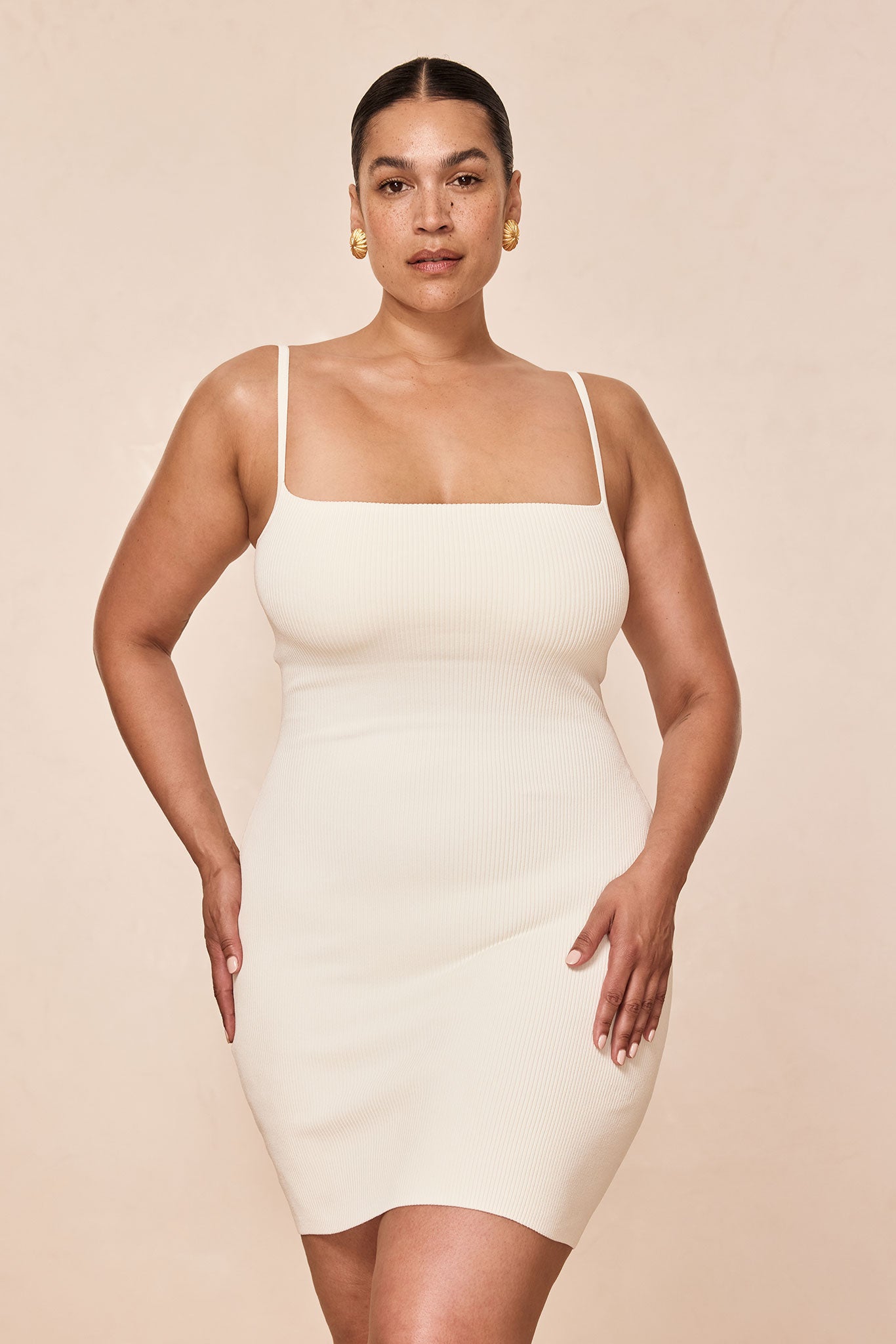 Model wearing ribbed ivory square-neck bodycon dress with thin straps and gold earrings, hands on hips, neutral background.