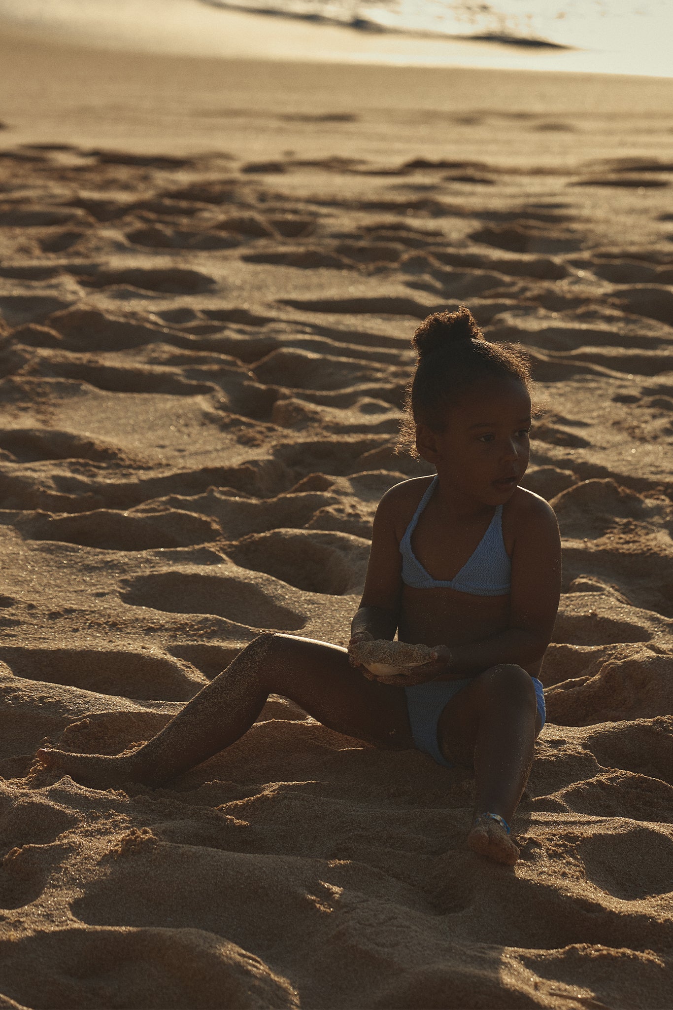 Young child in blue swimsuit sitting on sandy beach at sunset, holding a handful of sand