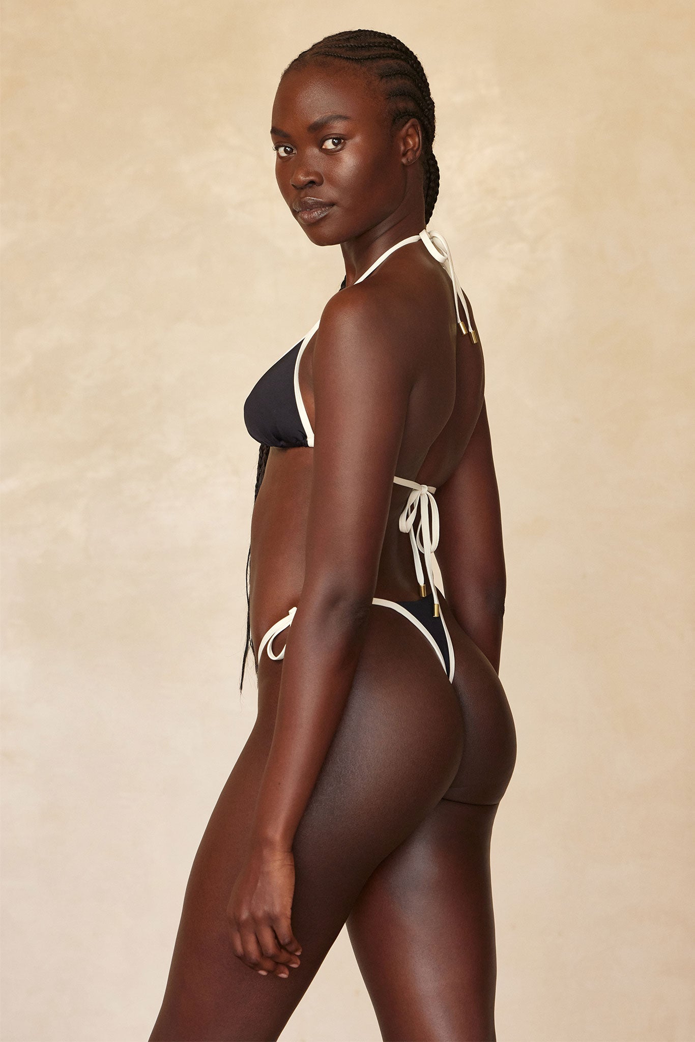 Dark-skinned woman in black string bikini with contrasting white trim, standing three-quarter back pose looking over shoulder