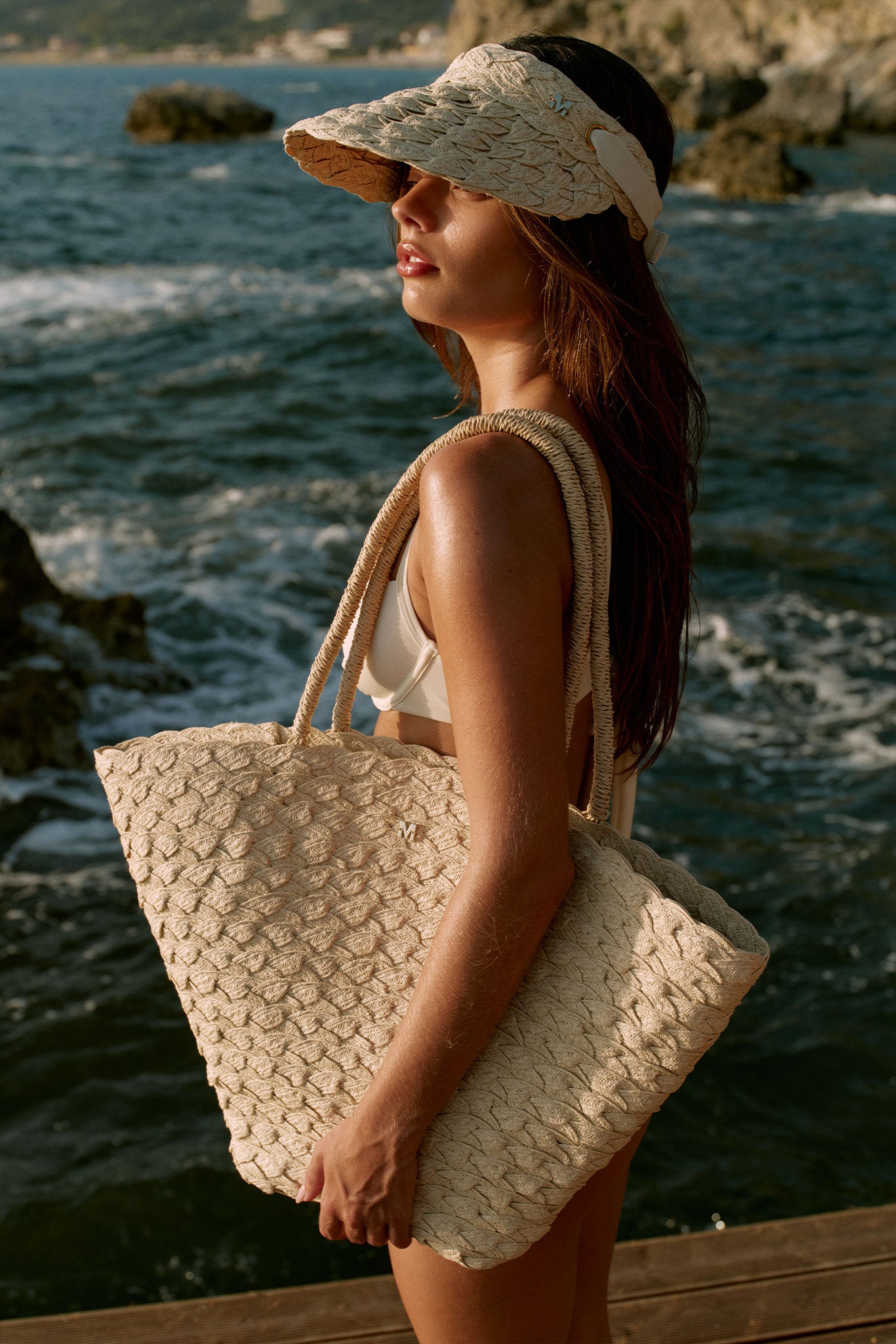 Young woman in bikini wearing woven visor, holding a large woven straw tote with M charm, standing on a seaside pier.