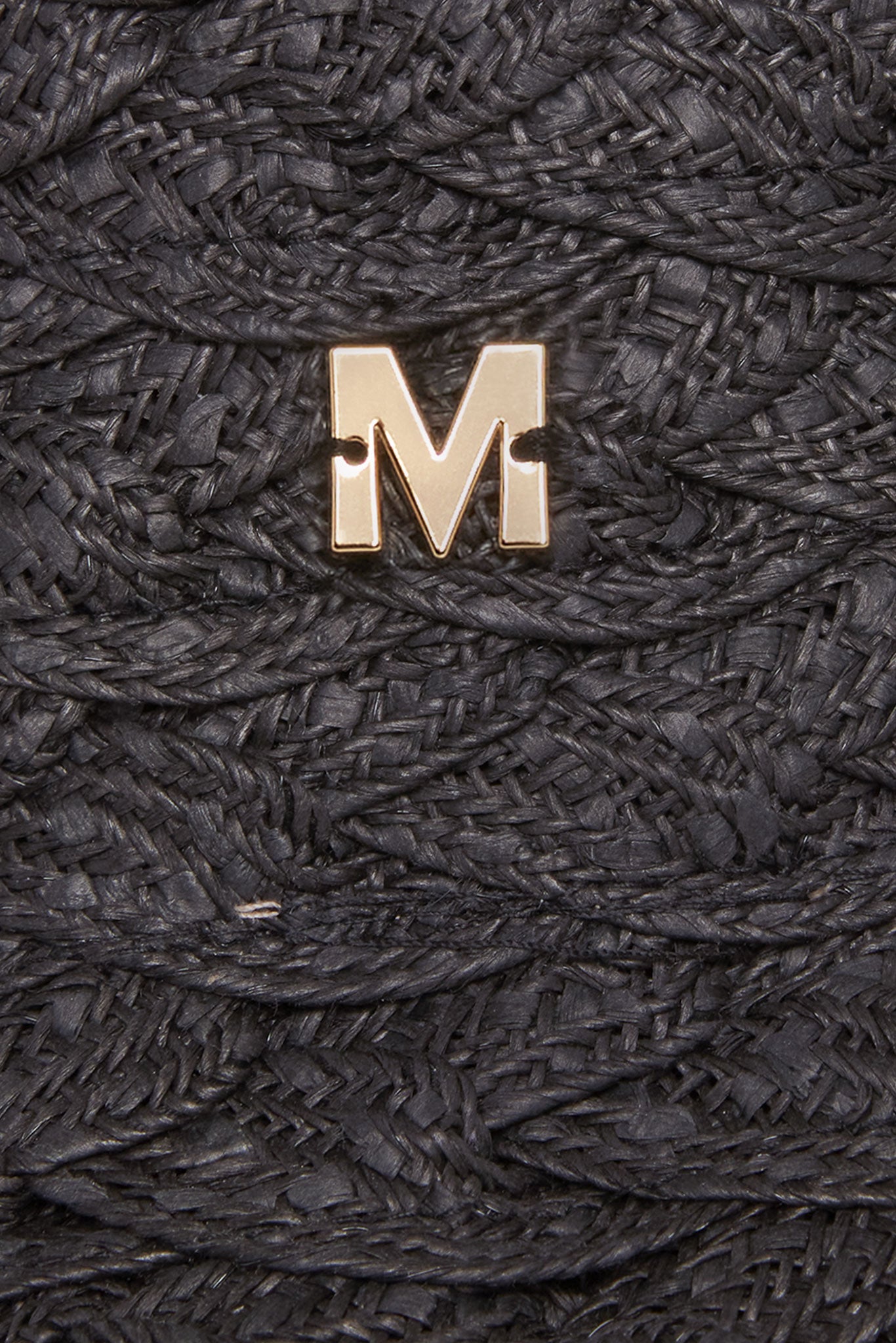 Gold metal M badge pinned to black braided straw fabric