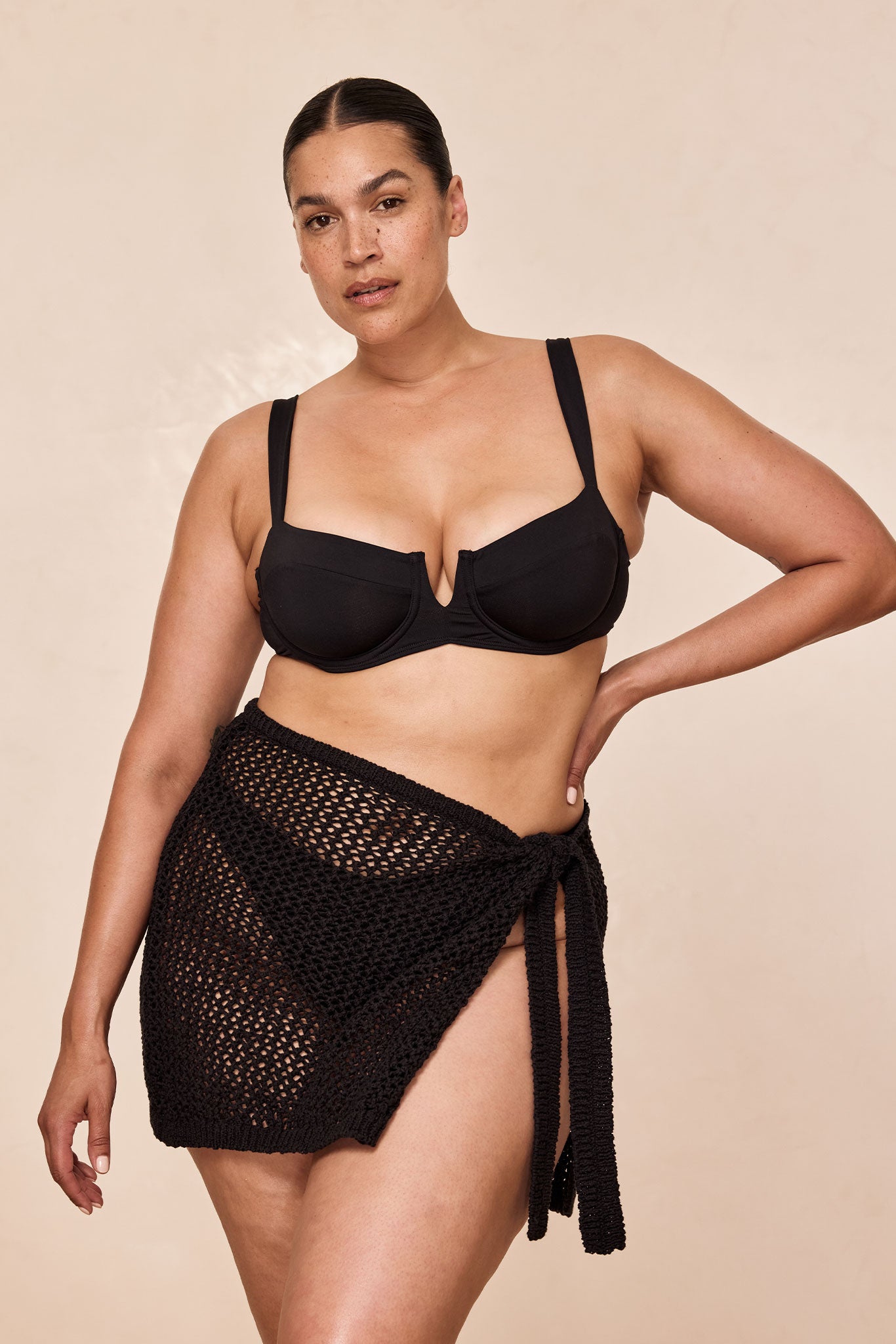 Model wearing black underwire bikini top and black crocheted wrap skirt, hand on hip against neutral backdrop.