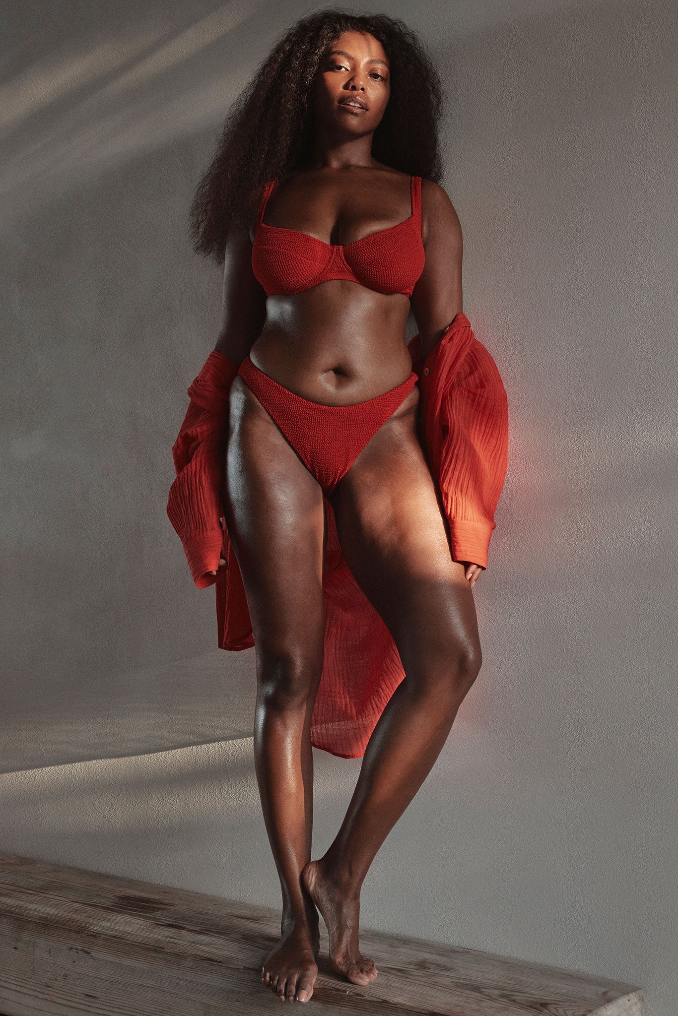 Plus-size model standing barefoot on a wooden platform wearing a textured red bikini and open red shirt.