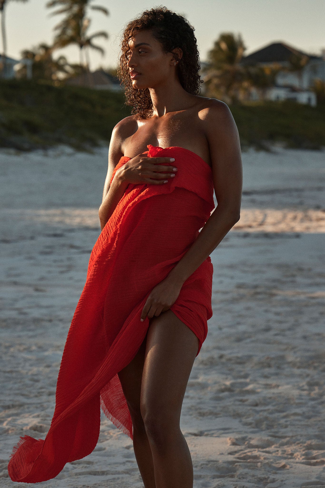Woman standing on sandy beach at sunset, wearing and holding a red wrap across her chest, looking left.