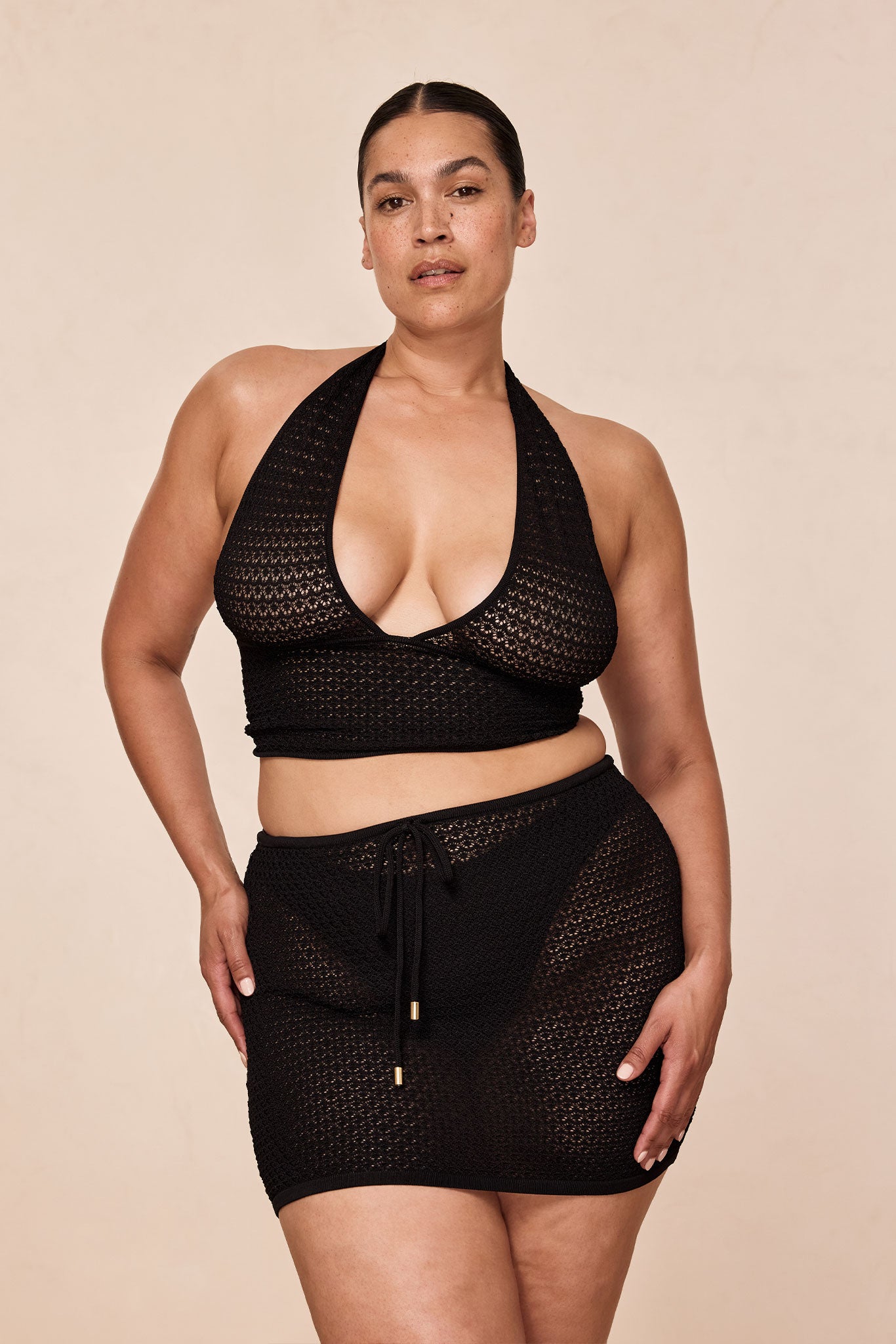 Plus-size woman wearing black crochet halter crop top and matching crochet mini skirt, standing and facing camera with hands at hips.