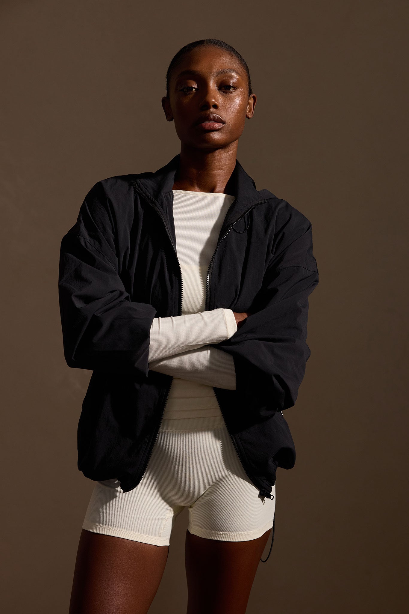 Adult woman standing with arms crossed wearing a black zip jacket over cream ribbed athletic top and shorts, studio portrait.