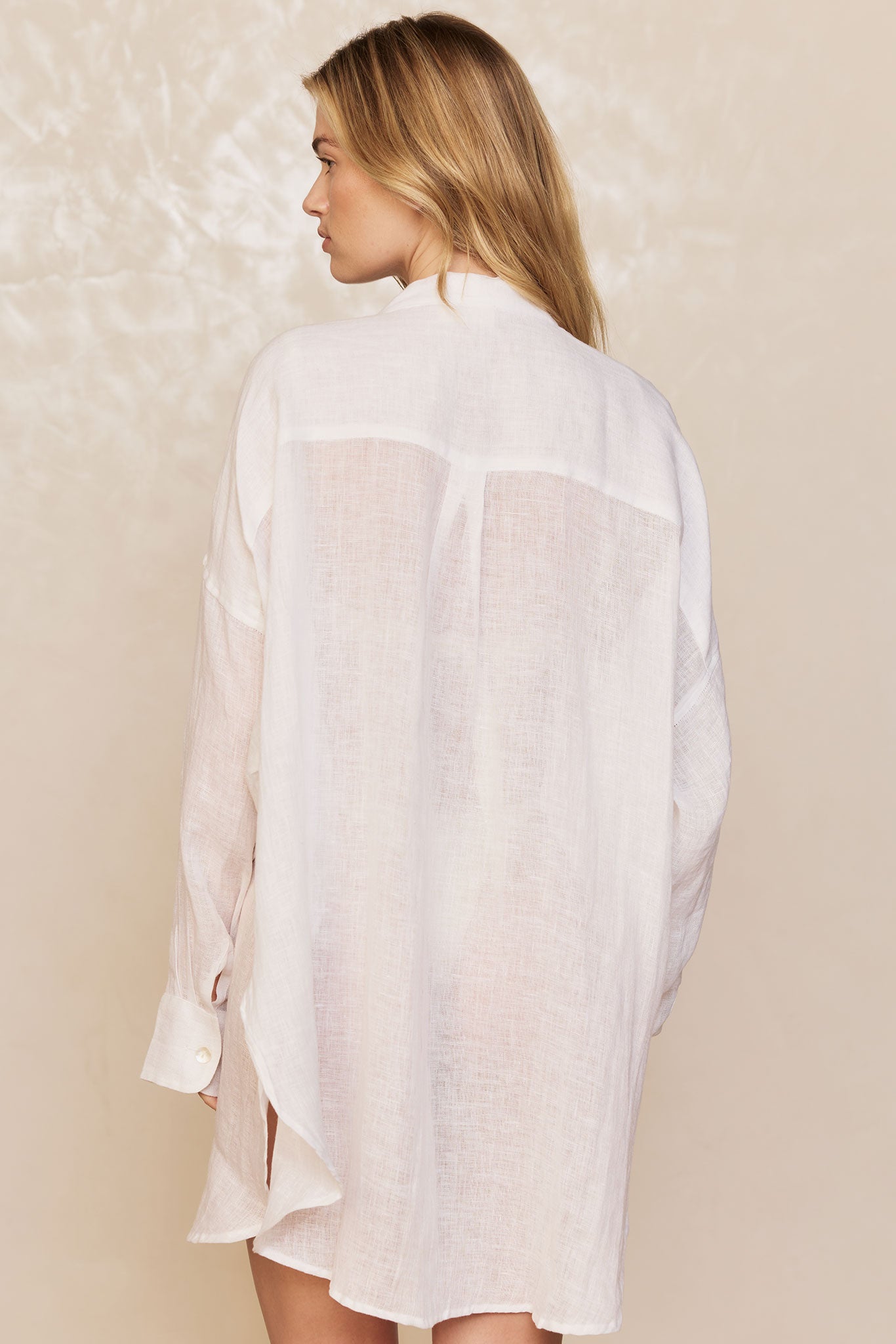 Back view of model wearing a sheer white linen long shirt with buttoned cuff and center back yoke.