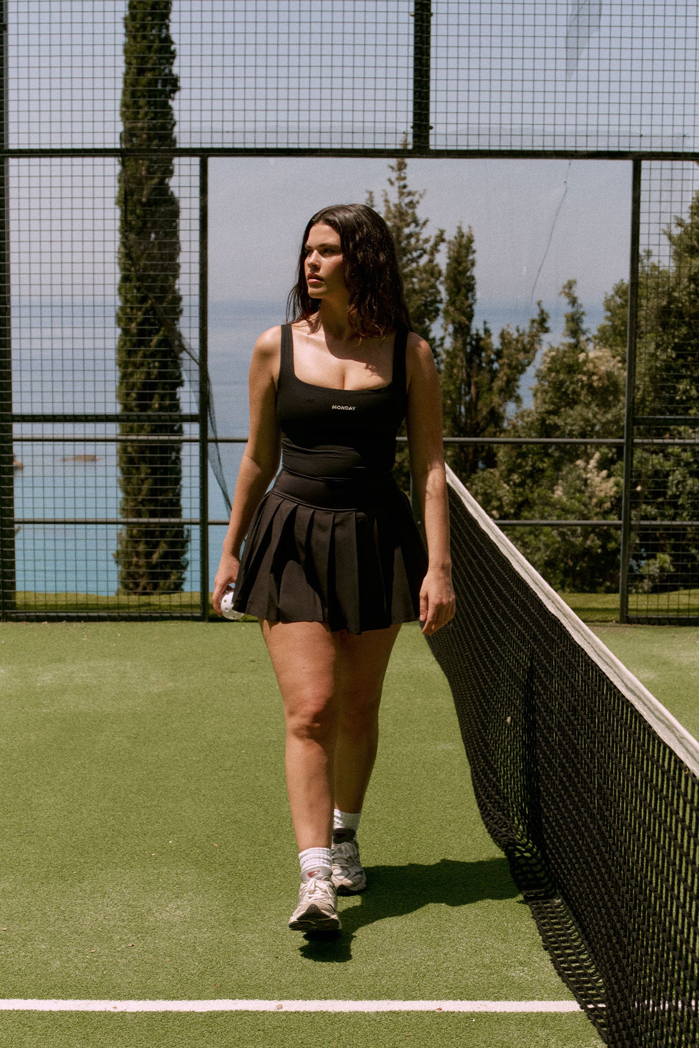 Woman walking along a tennis net on a court, wearing a black pleated skirt and top labeled 'MONDAY'.