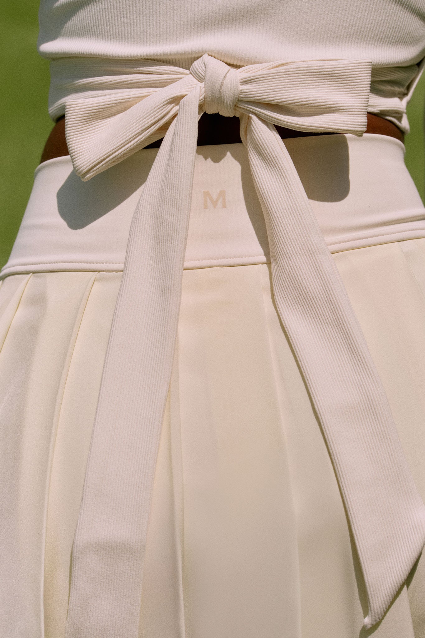 Cream pleated skirt waistband with wide ribbed bow tied at back; small M logo centered on the waistband