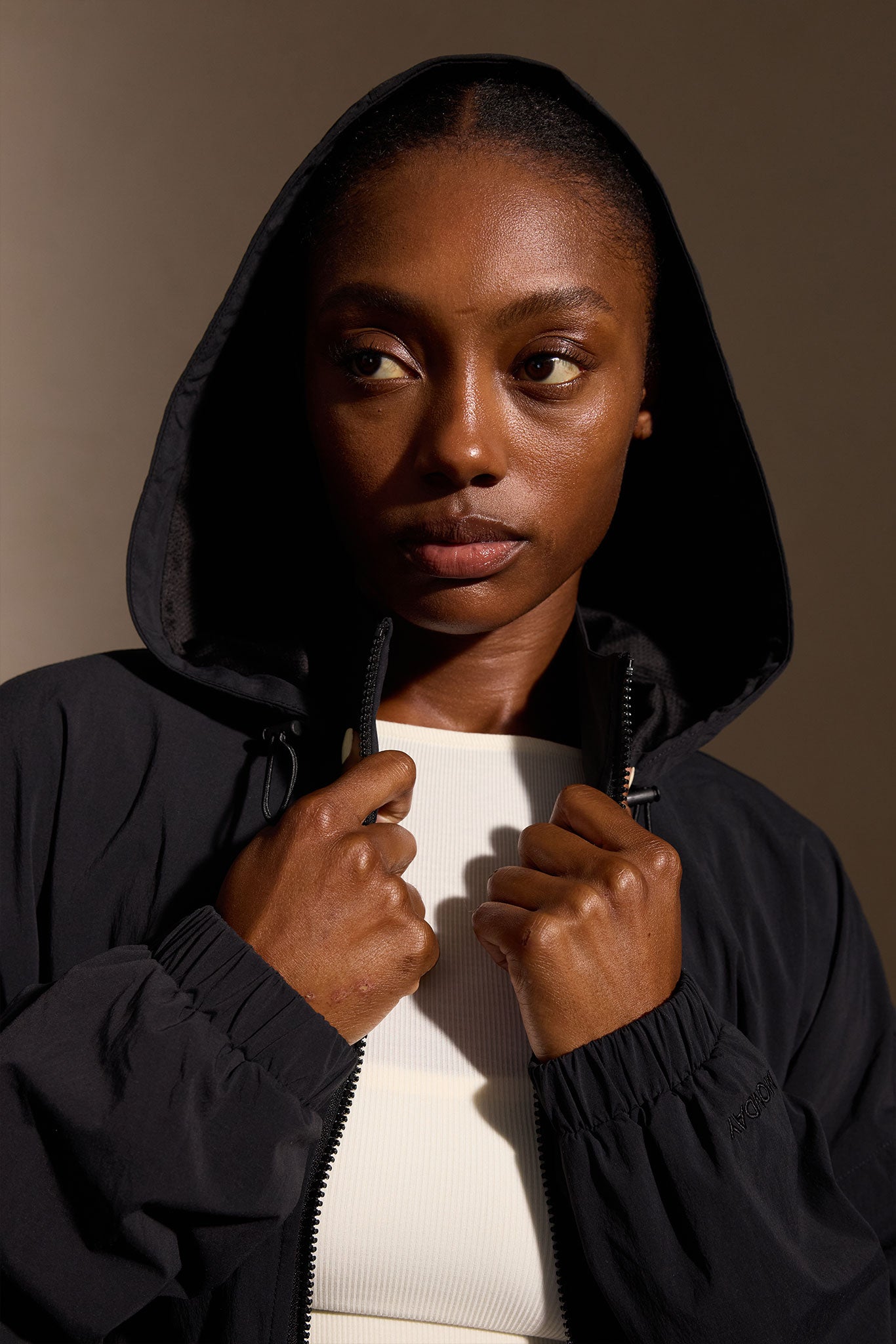 Woman wearing a black hooded jacket, holding the zipper at her chest and looking slightly to the left against a neutral background.