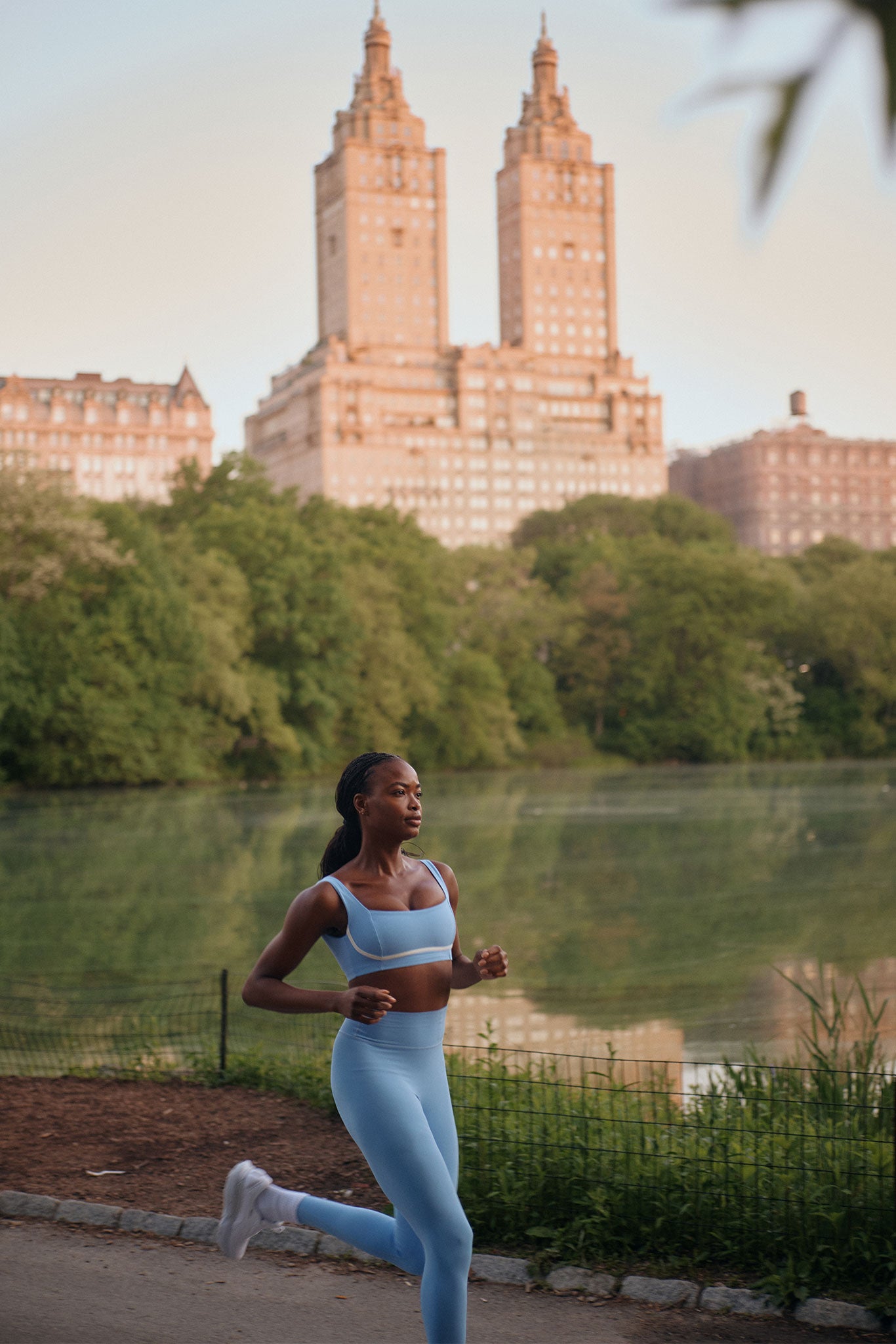 Woman jogging in blue activewear on a park path by a lake, with trees and twin-towered apartment building in the background.