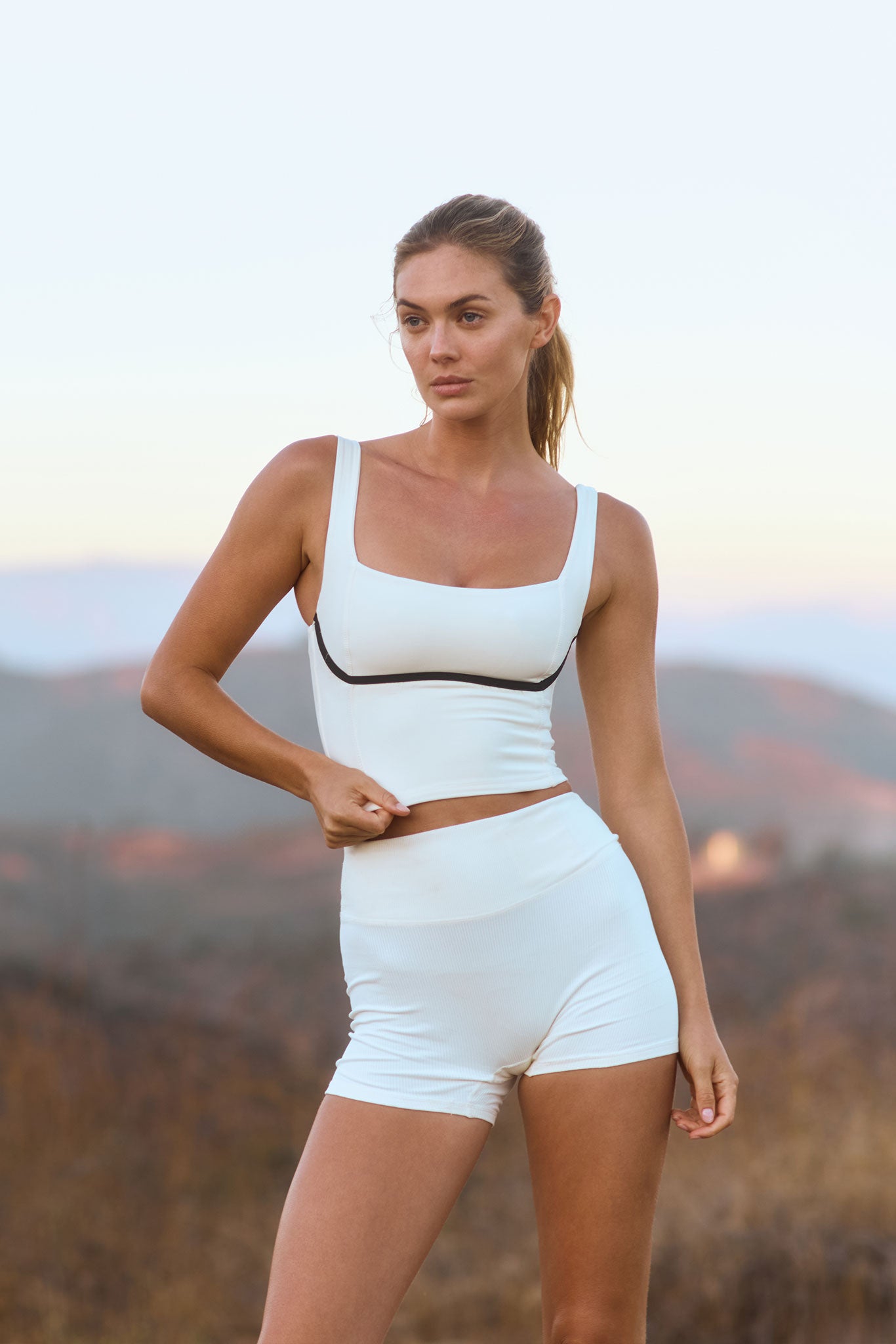 Woman in white cropped athletic tank and high-waisted shorts standing outdoors at dusk, pulling at her top