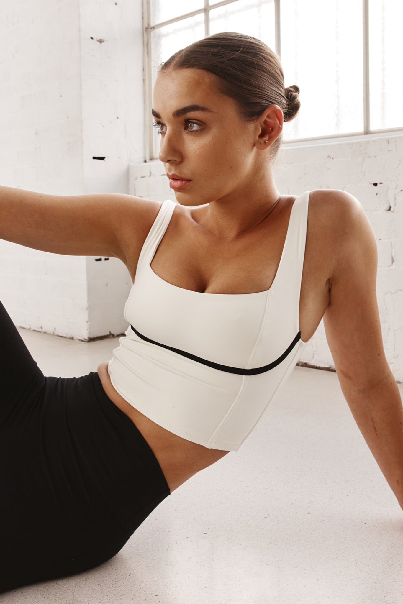 Woman reclining on floor in white cropped tank top with black trim and black leggings, looking left near large studio window.