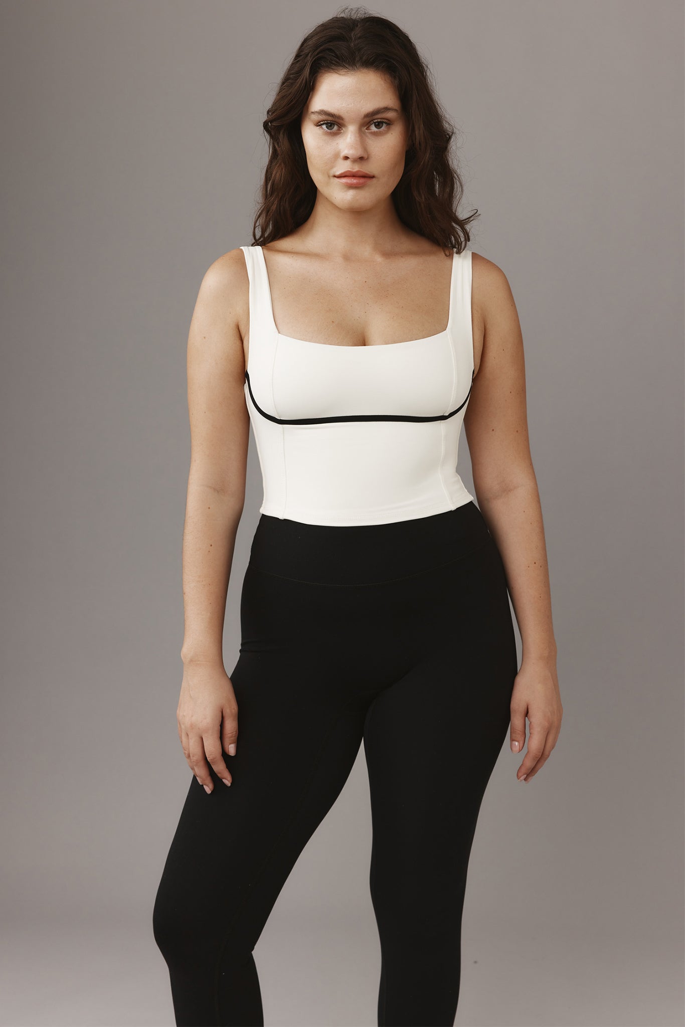 Woman standing wearing white cropped square-neck tank top with black piping and high-waist black leggings on gray background.