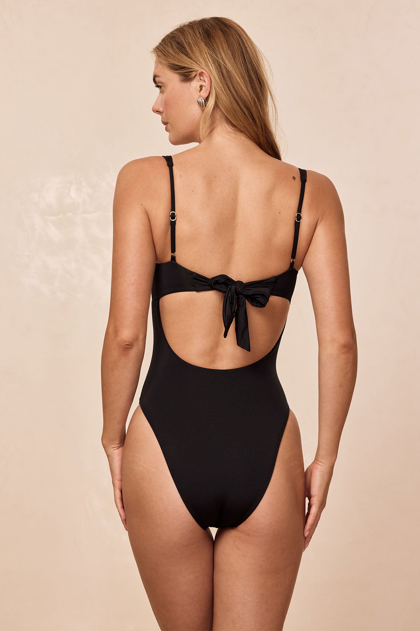 Woman shown from back wearing a black one-piece swimsuit with adjustable straps, tied bow back, and high-cut leg openings.