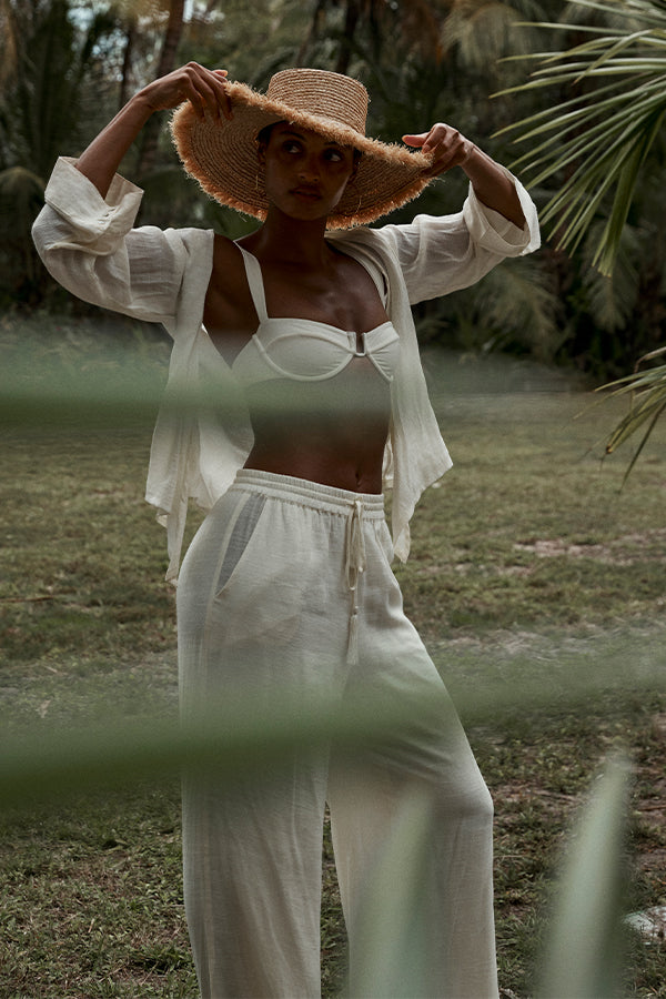Woman in white linen pants, top, and straw sun hat adjusting the hat in a tropical garden